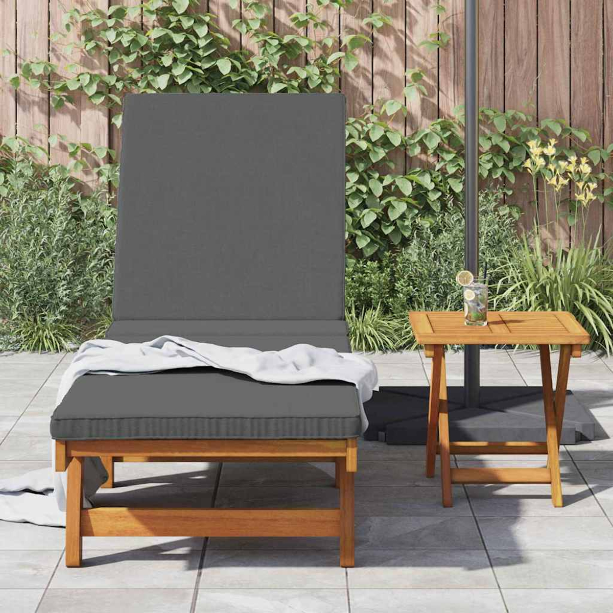 Graue Outdoor-Liege mit weißer Decke und kleinem Holztisch mit Getränk auf einer Terrasse. Grüne Pflanzen im Hintergrund.