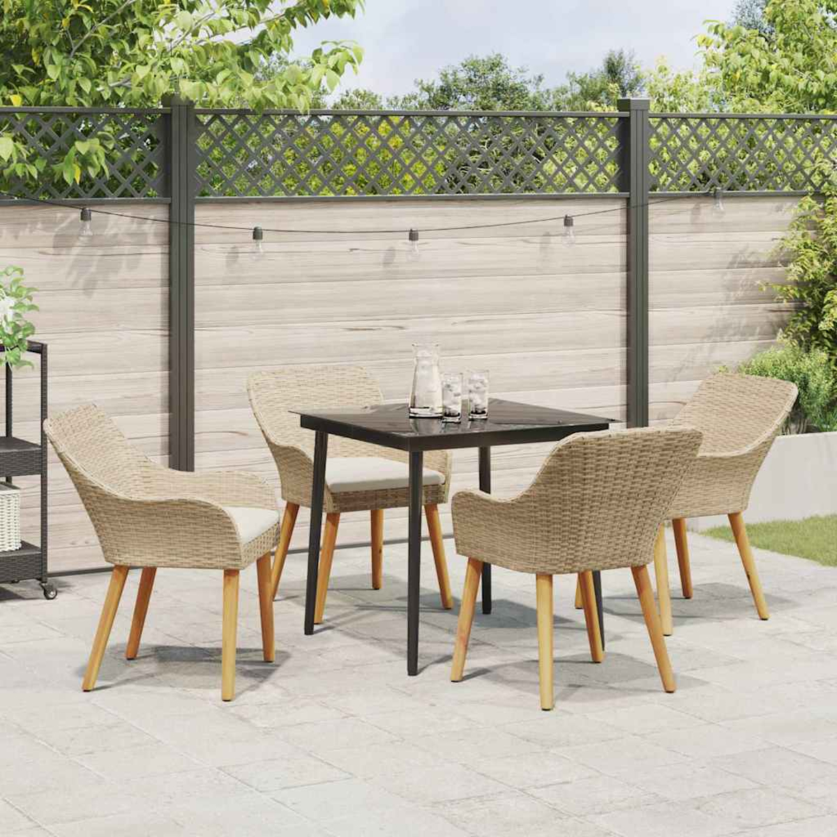 Essgarnitur im Freien: quadratischer schwarzer Tisch, vier Korbstühle, auf einer Terrasse. Hintergrund: Holzzaun, Grün und Lichterketten.