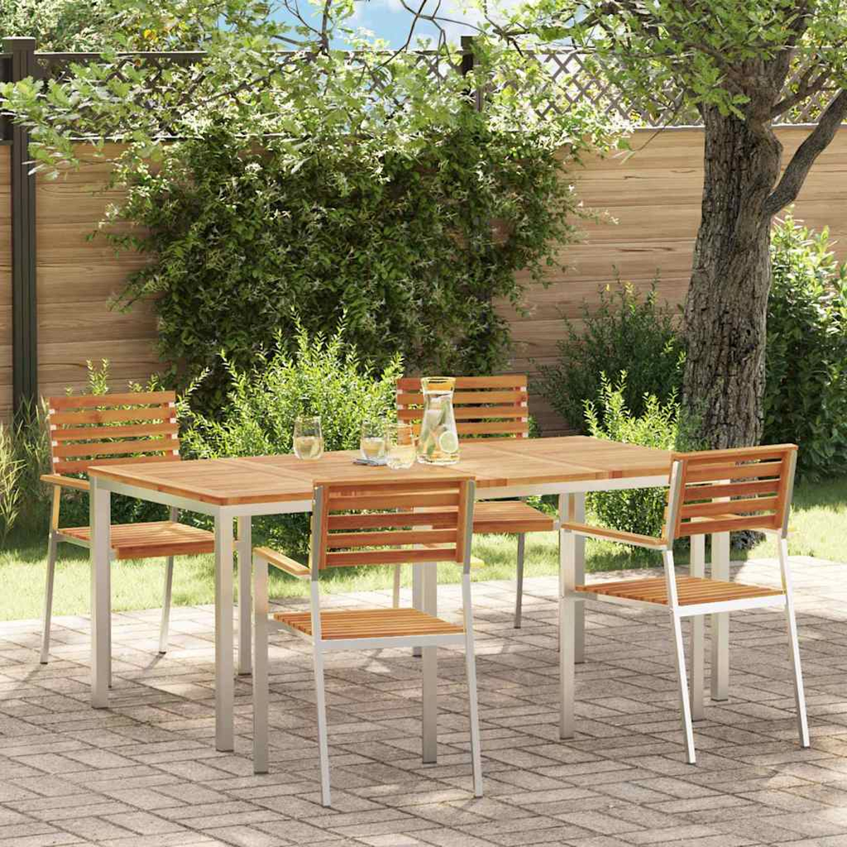Ein Holztisch und Stühle auf einer Terrasse. Auf dem Tisch stehen ein Wasserkrug und Gläser. Grünes Laub bildet den Hintergrund.