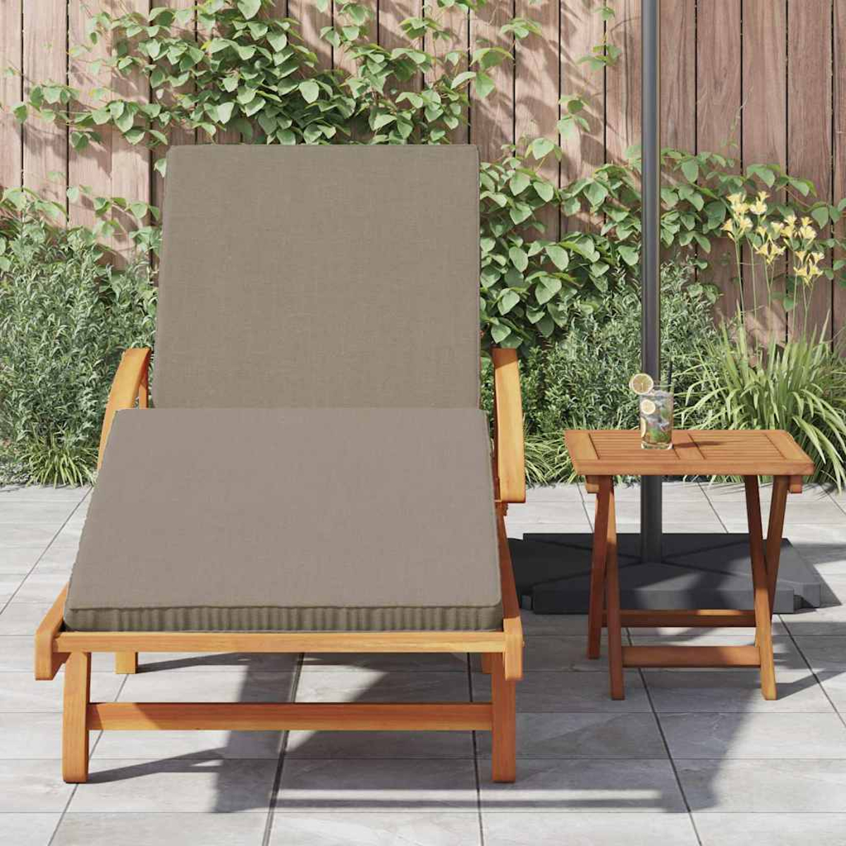 Eine beige gepolsterte Liege und ein kleiner hölzerner Beistelltisch stehen auf einer Terrasse. Grüne Pflanzen und ein Holzzaun befinden sich im Hintergrund.