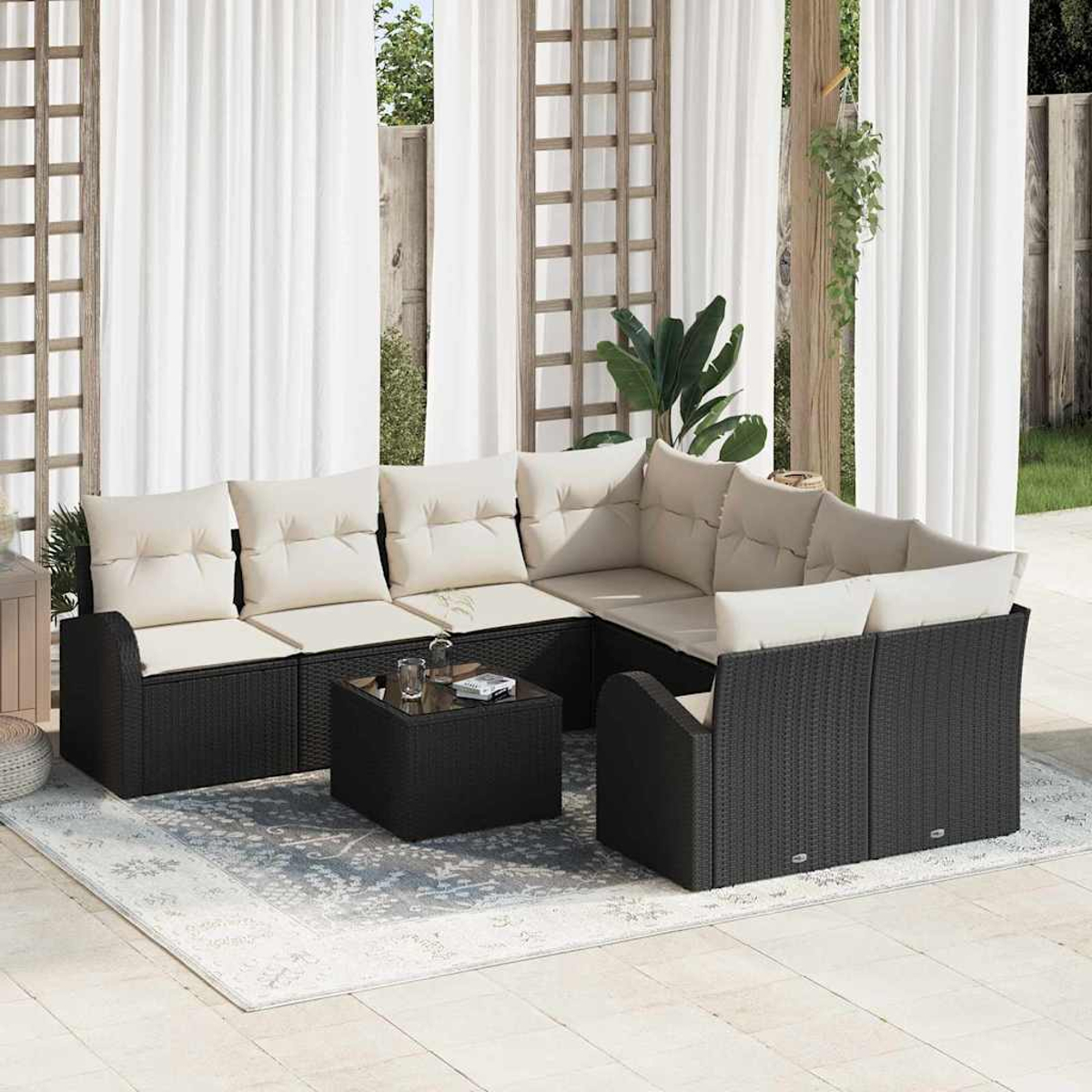 Sofá seccional para exteriores con cojines beige, estructura de mimbre negro y mesa de centro de vidrio. Cortinas blancas y plantas.
