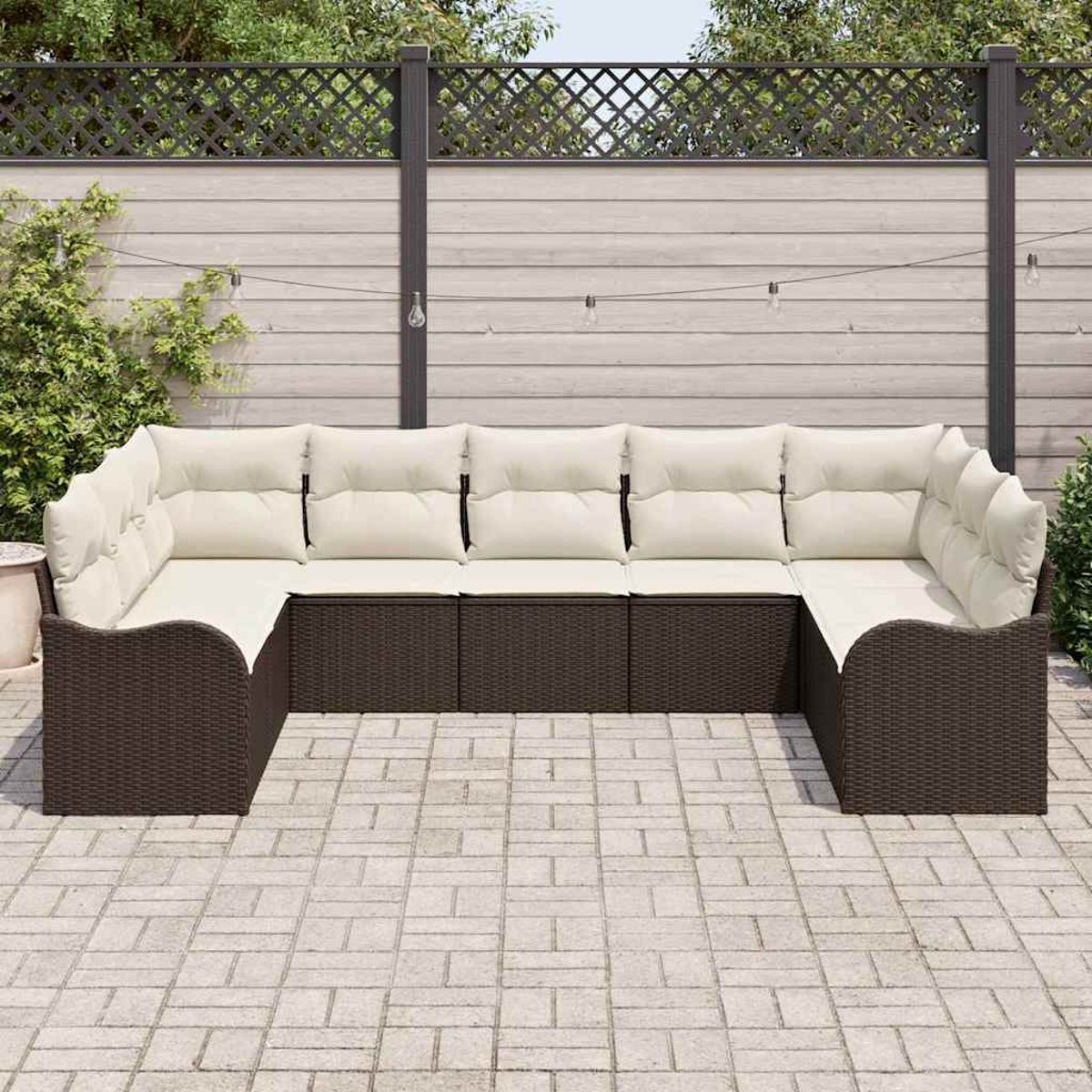 Outdoor-Ecksofa mit cremefarbenen Kissen. Braune Korbbasis auf einer Ziegelterrasse. Holzzaun und Grün in Hintergrund.