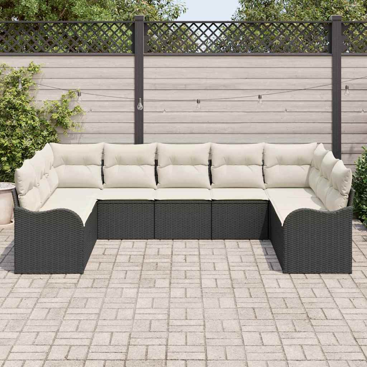Ein graues Korbsessel-Ecksofa mit cremefarbenen Kissen steht auf einer Ziegelsteinterrasse. Ein Holzzaun und Grünzeug sind im Hintergrund.