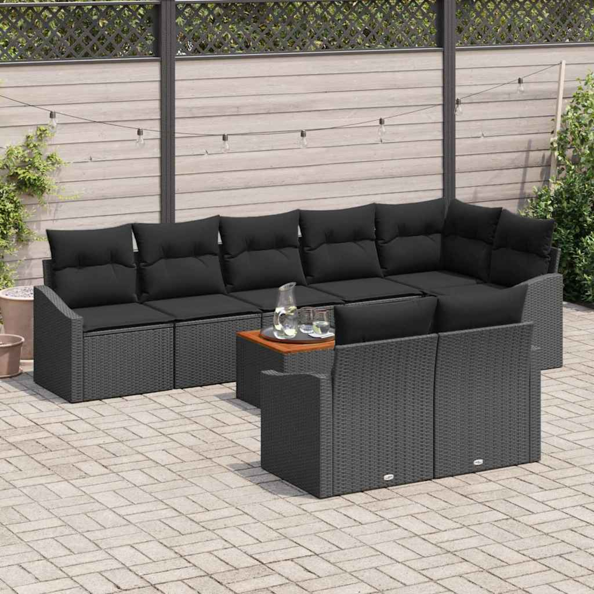 Juego de patio al aire libre con cojines negros, muebles de mimbre grises y una mesa de café de madera en un patio de ladrillos. Valla de fondo.