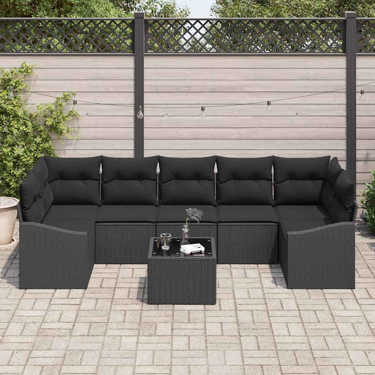 Outdoor-Ecksofa mit schwarzen Kissen und einem quadratischen Tisch. Auf einer Ziegelterrasse vor einem Holzzaun.