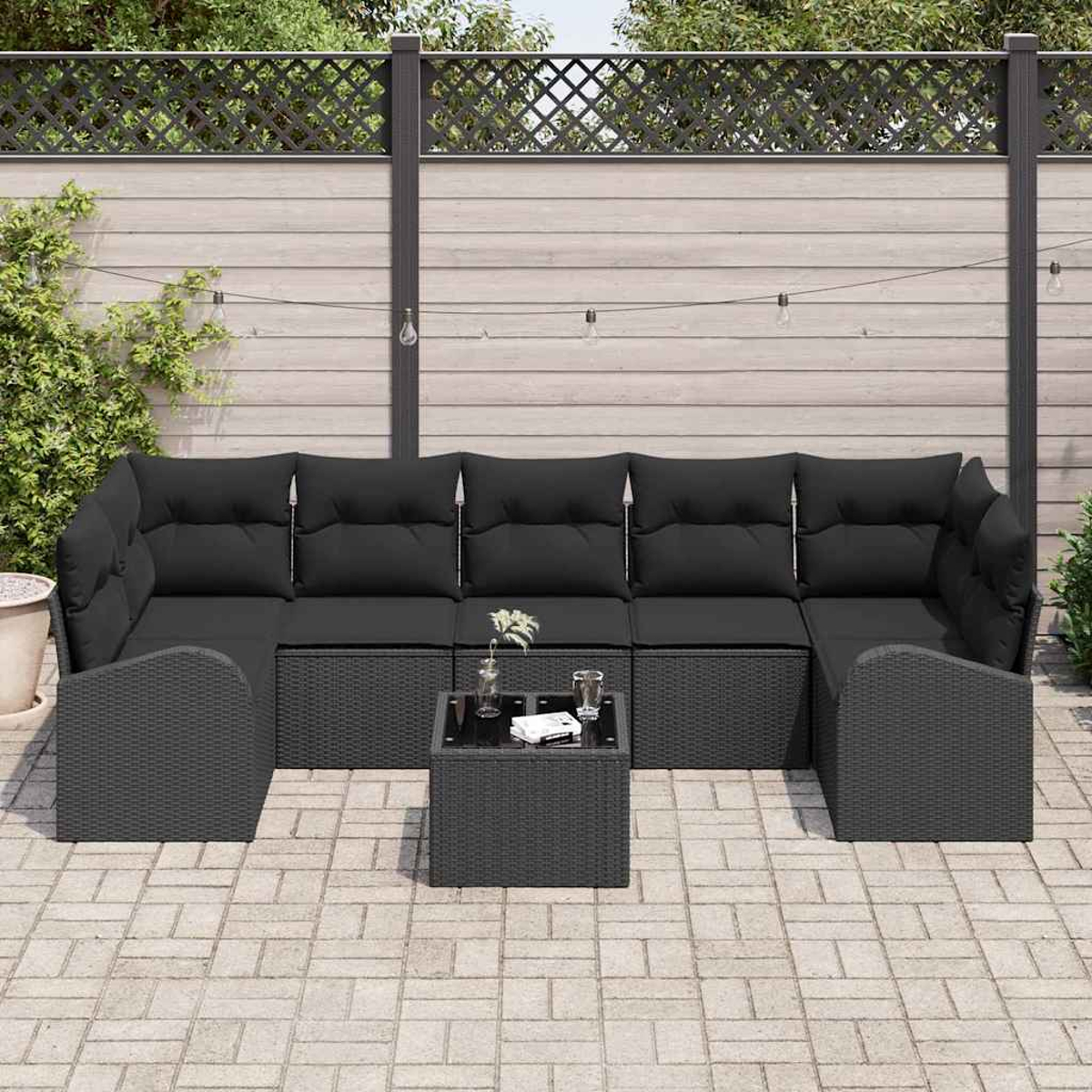 Schwarzes Outdoor-Wicker-Ecksofa mit schwarzen Kissen und einem kleinen Tisch. Der Hintergrund umfasst einen Holzzaun und Pflanzen.
