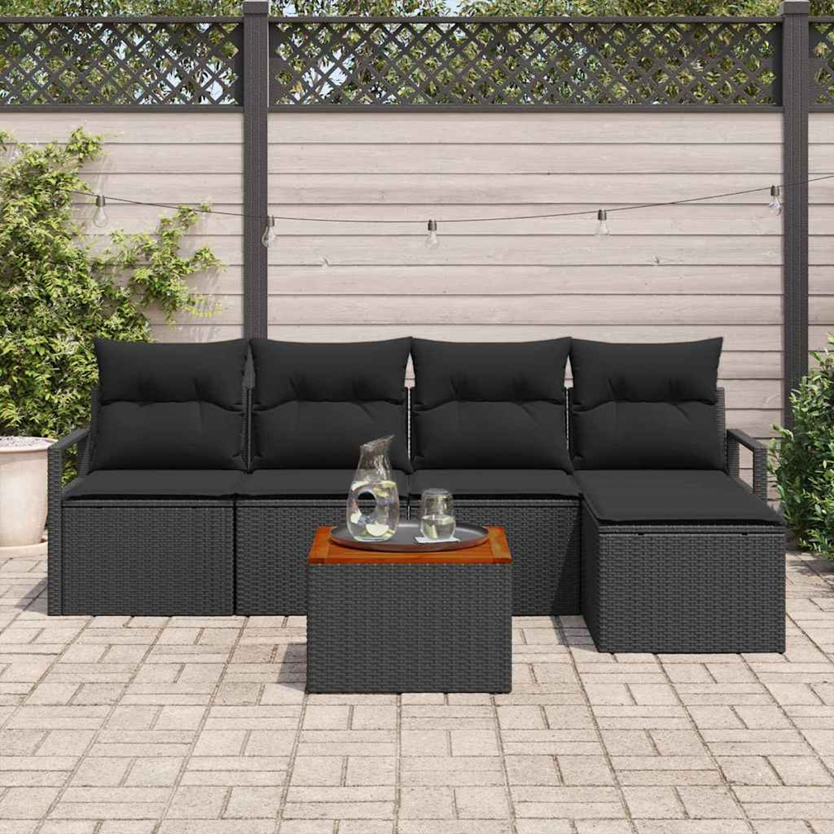 Garten-Sitzgruppe mit schwarzen Kissen und einem kleinen Tisch mit Tablett. Ein Zaun und Pflanzen sind im Hintergrund.