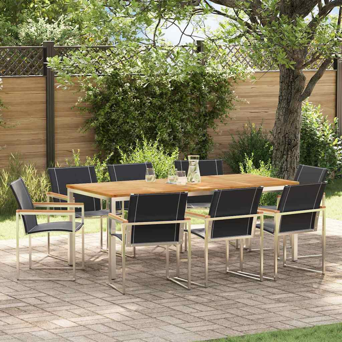 Juego de comedor al aire libre: una mesa de madera con ocho sillas negras, sobre un patio de ladrillos, rodeado de vegetación y una valla de madera.