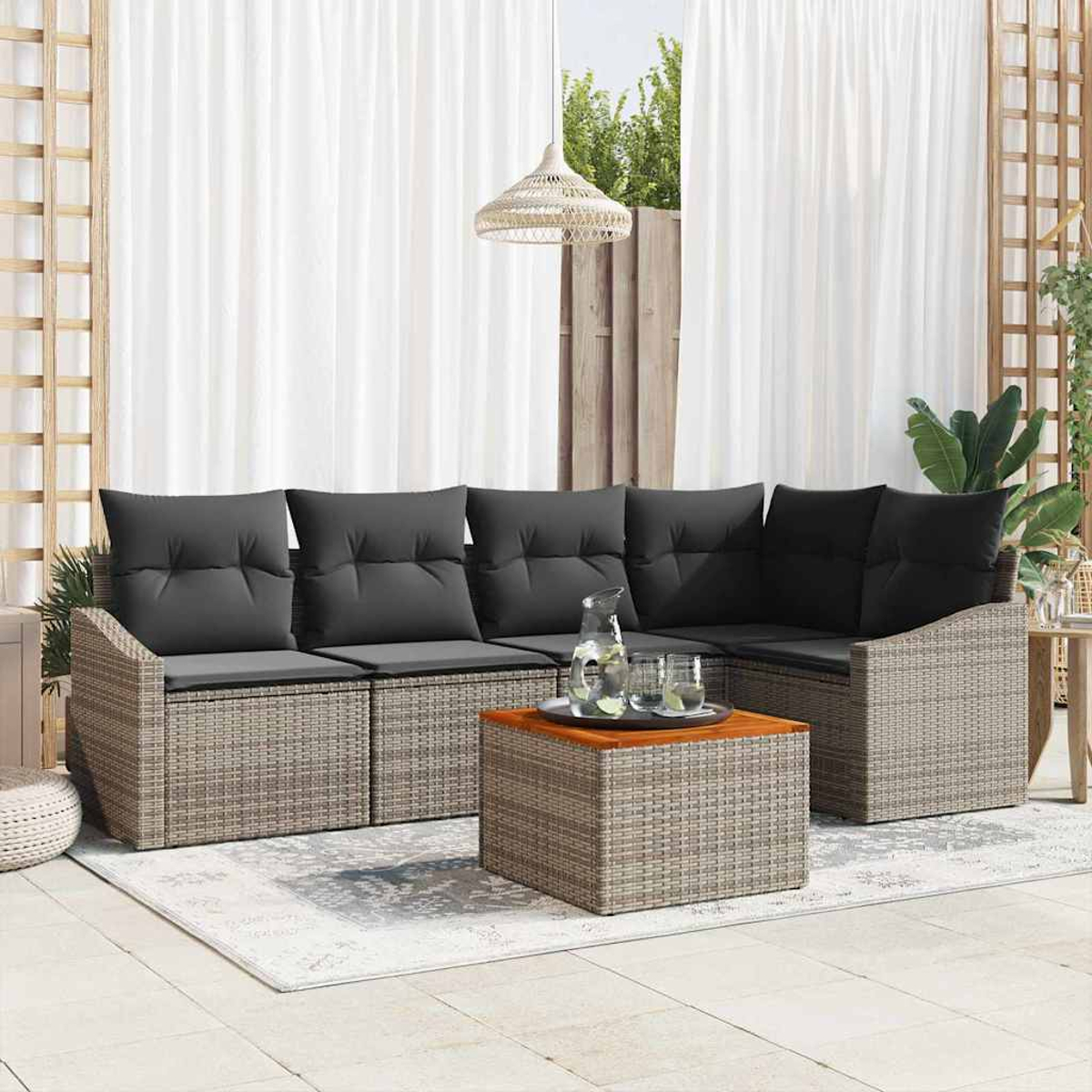 Geflochtene Terrassenmöbel mit grauen Kissen. Ein quadratischer Couchtisch trägt ein Tablett mit Gläsern und einer Wasserkaraffe. Weiße Vorhänge.
