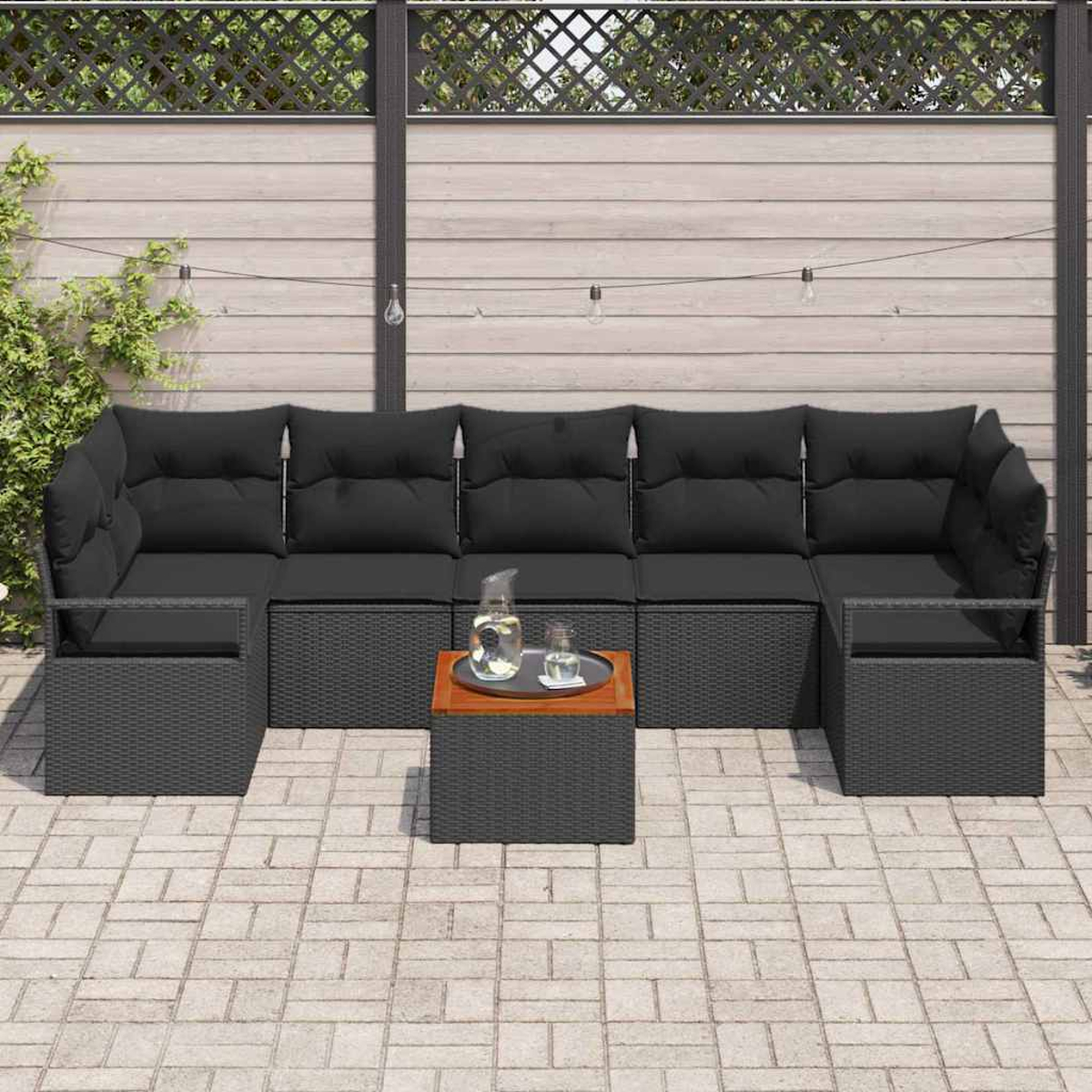 Sofá seccional para exteriores con cojines negros y una mesa pequeña con bebidas. Situado en un patio de ladrillo con valla de madera al fondo.