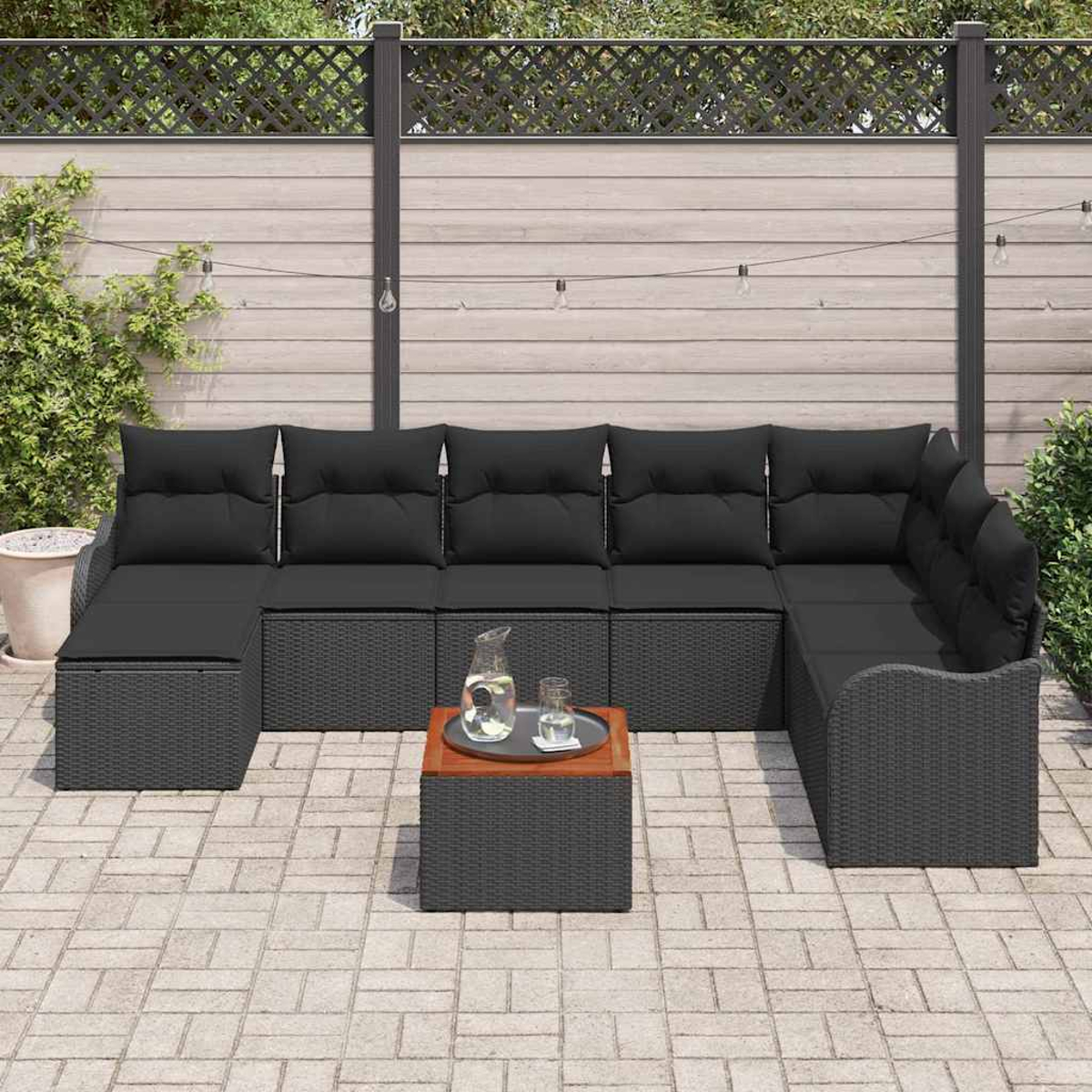 Outdoorsofa mit schwarzen Kissen und einem kleinen Tisch auf einer Terrasse. Im Hintergrund ein Zaun, Grün und Lichterketten.