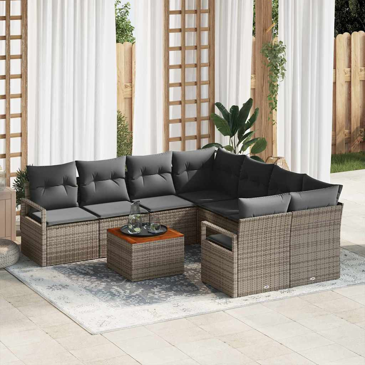Outdoor-Ecksofa mit grauen Kissen und einem kleinen Tisch auf einem gemusterten Teppich. Weiße Vorhänge und Holzzaun im Hintergrund.