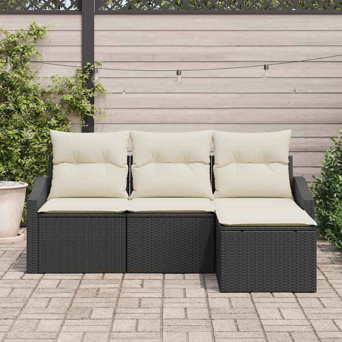 Ein schwarzes Outdoor-Sofa aus Korbgeflecht mit cremefarbenen Kissen auf einer Ziegelterrasse, vor einem Holzzaun mit Lichterketten.