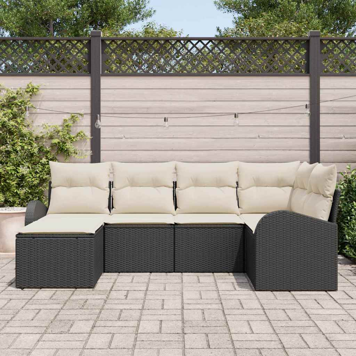 Ein schwarzes Korbsessel-Sofa mit cremefarbenen Kissen steht auf einer Terrasse. Im Hintergrund befindet sich ein Holzzaun mit Grün.