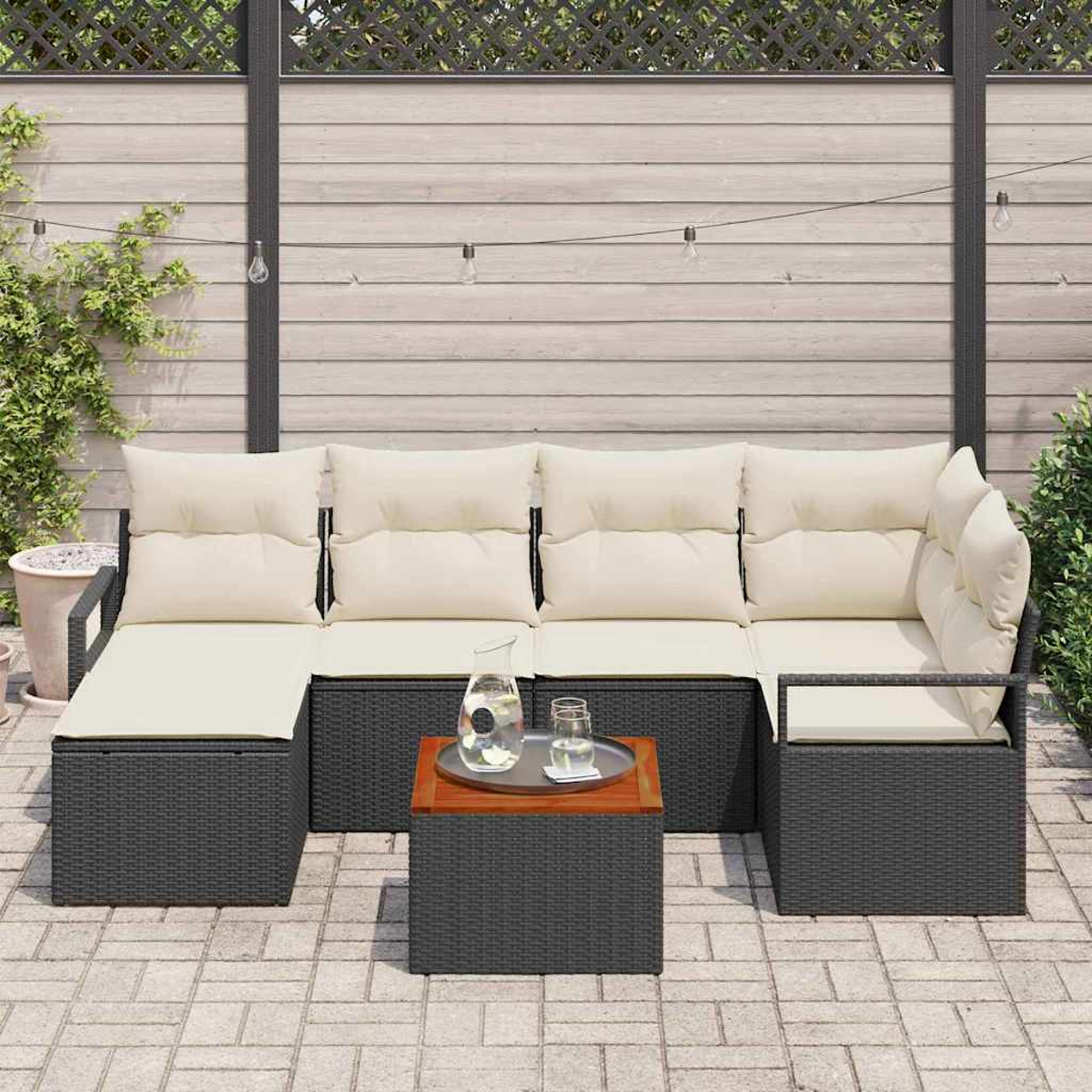 Garten-Sitzgruppe mit beigen Kissen, dunkelgrauem Korbgeflecht und einem kleinen Holztisch mit Wasserkaraffe. Hintergrund ist Holzzaun.