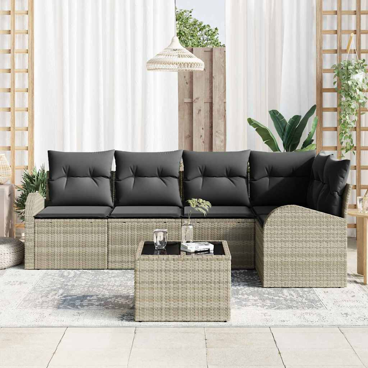 Outdoor-Rattansofa mit grauen Kissen, Couchtisch und transparenten Vorhängen. Pflanzen und Hängeleuchte vervollständigen die Szene.