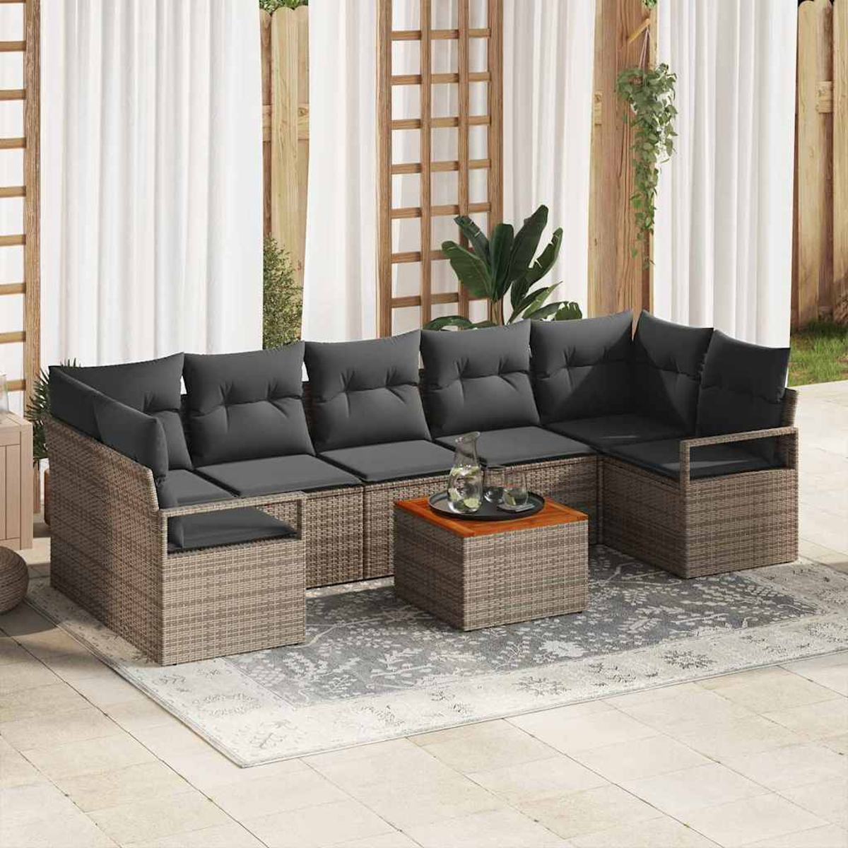 Ein Ecksofa im Freien mit grauen Kissen und einem kleinen Tisch steht auf einem Teppich, Vorhänge im Hintergrund. Pflanzen und Holzstrukturen umgeben.