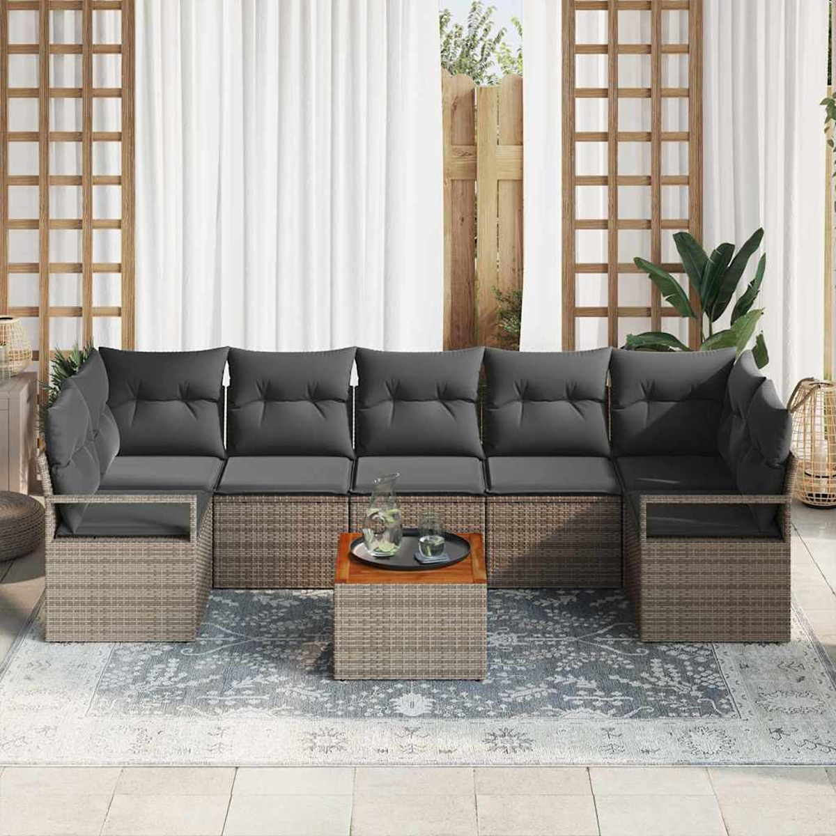 Outdoor-Sitzarrangement mit grauen Kissen, einem Holztisch und einem gemusterten Teppich. Weiße Vorhänge und Holzgitter im Hintergrund.