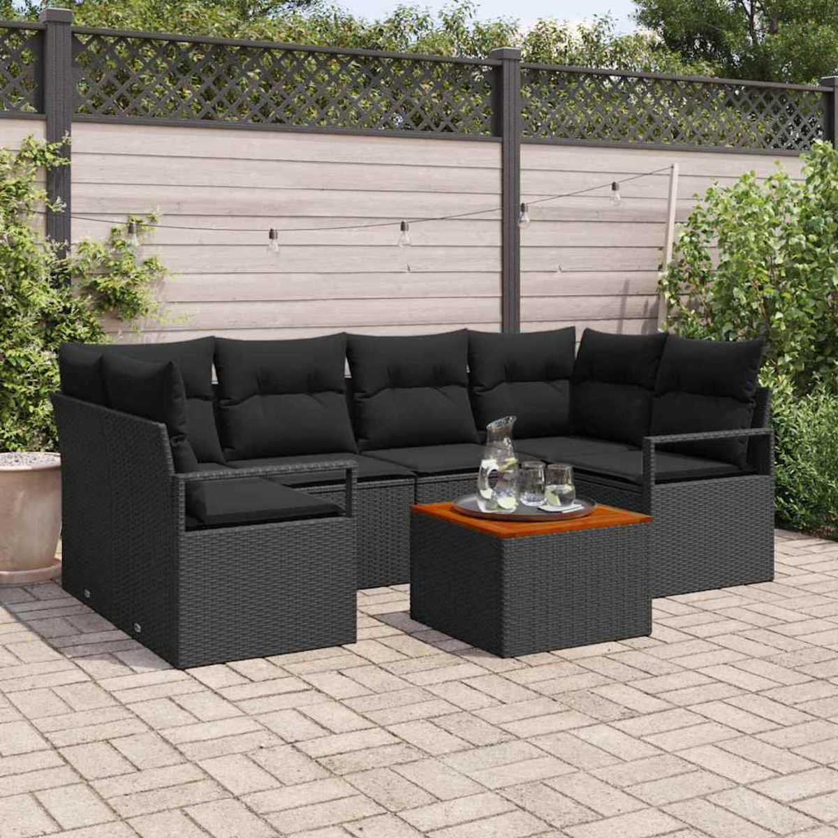 Schwarzes Outdoor-Wicker-Sofaset mit schwarzen Kissen und einem hölzernen Couchtisch. Dahinter ein Holzzaun, Pflanzen und Lichterketten.