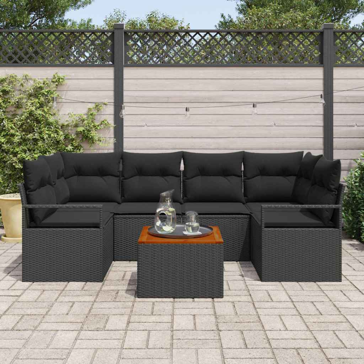 Garten-Terrassenset mit schwarzen Kissen auf grauen Korbmöbeln, einem Holztisch, einer Glaskanne und einem Zaun im Hintergrund.