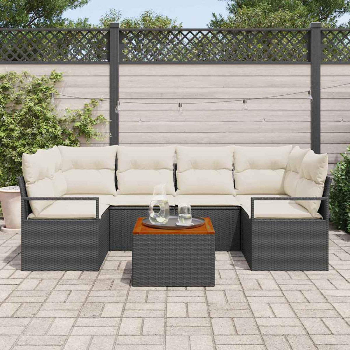 Garten-Sitzgruppe mit grauem Korbgeflecht, cremefarbenen Kissen und einem Holztisch. Auf einer Terrasse mit Zaun und Grün im Hintergrund.