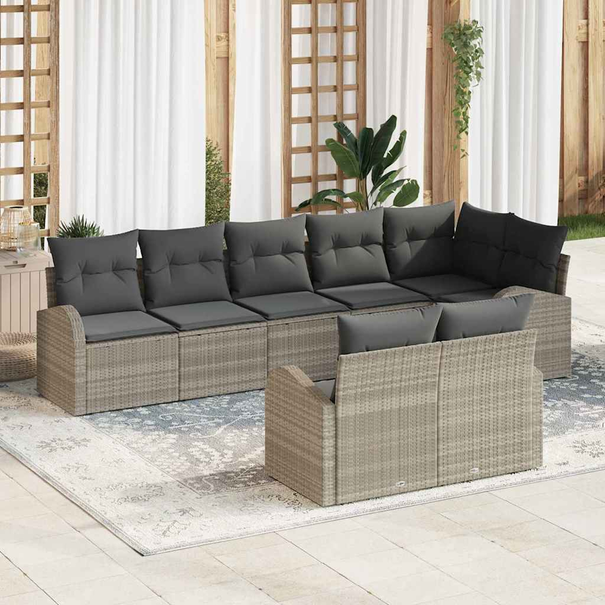 Rattan-Sofagarnitur mit grauen Kissen auf einer Terrasse. Im Hintergrund ein weißer Vorhang und Holzgitter. Grüne Pflanzen sind sichtbar.