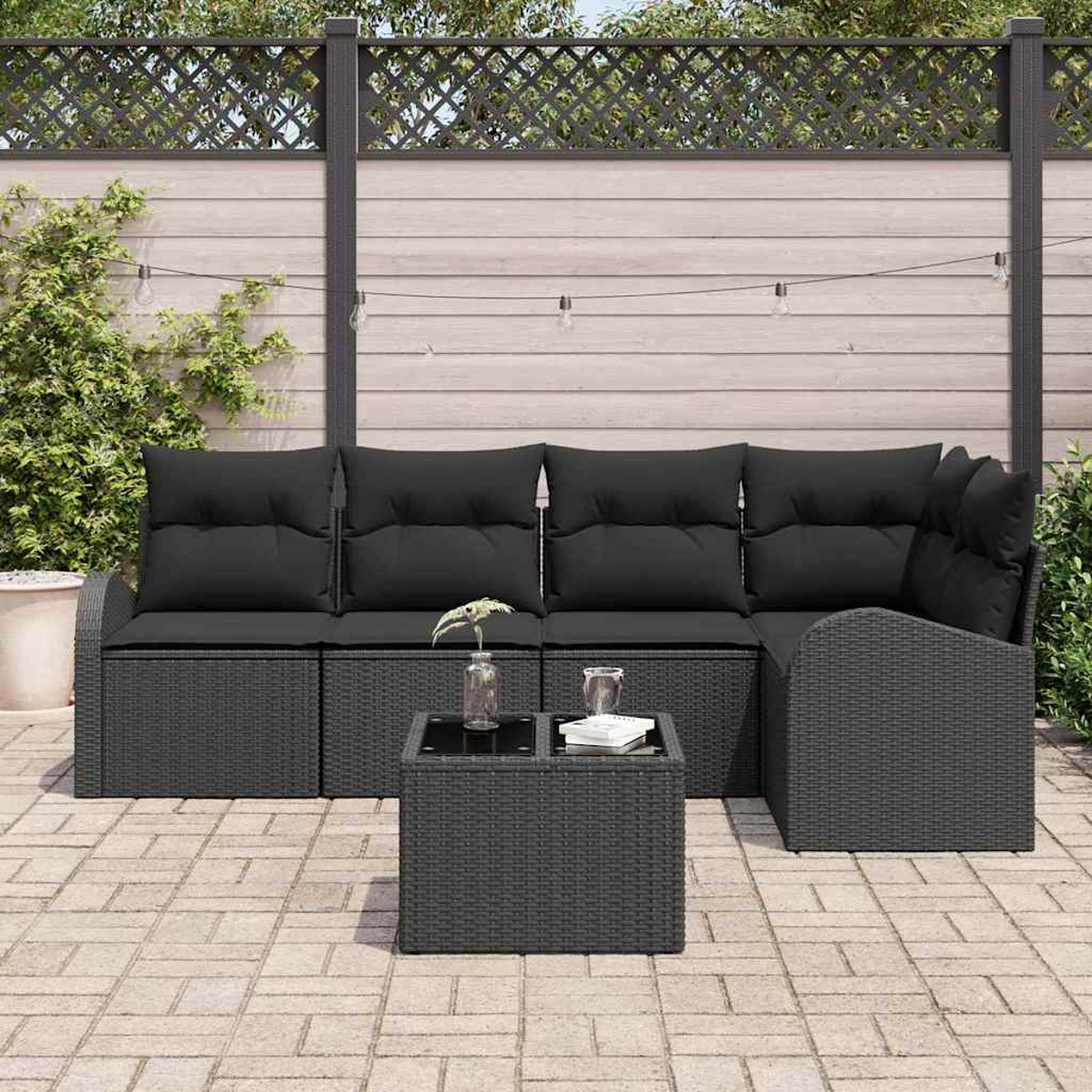Juego de patio exterior con cojines negros. Incluye sofá esquinero, mesa pequeña y luces, contra una valla de madera.