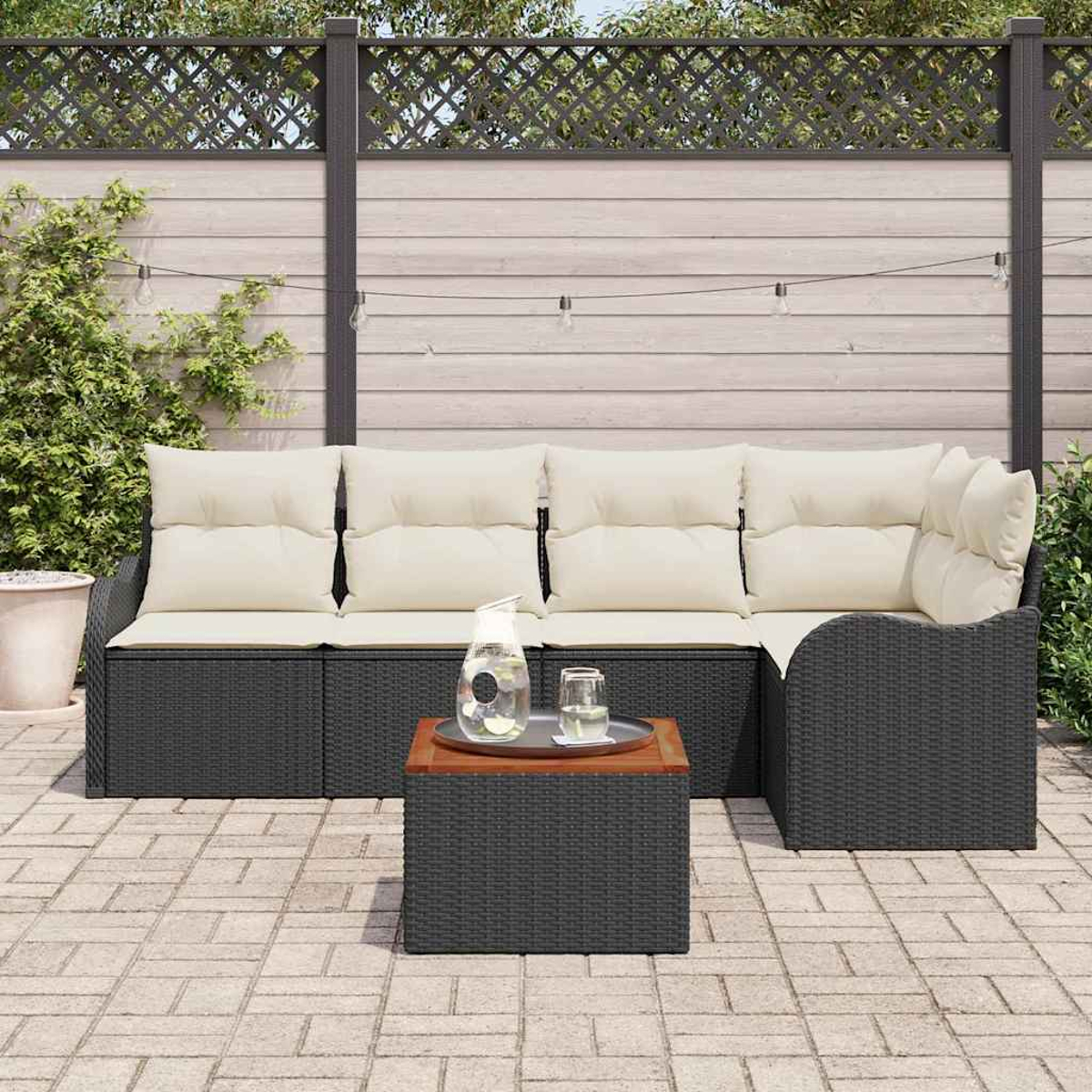 Ein Terrassenset mit einem schwarzen Korbmöbelsofa, beigen Kissen und einem Couchtisch mit Holzplatte. Im Hintergrund ein Zaun und Pflanzen.