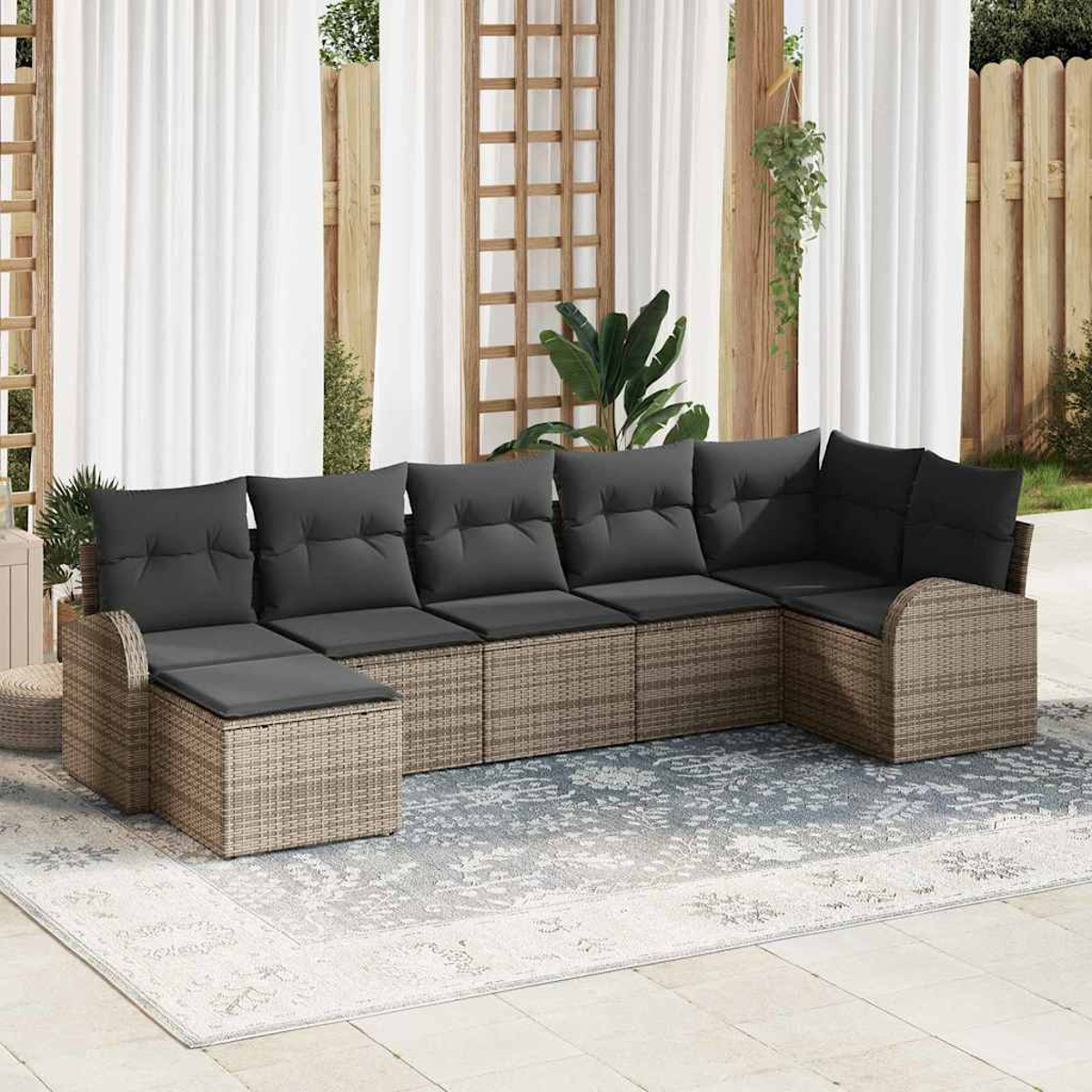 Ein Outdoor-Rattansofa mit grauen Kissen auf einem gemusterten Teppich, weißen Vorhängen und einem Holzzaun.