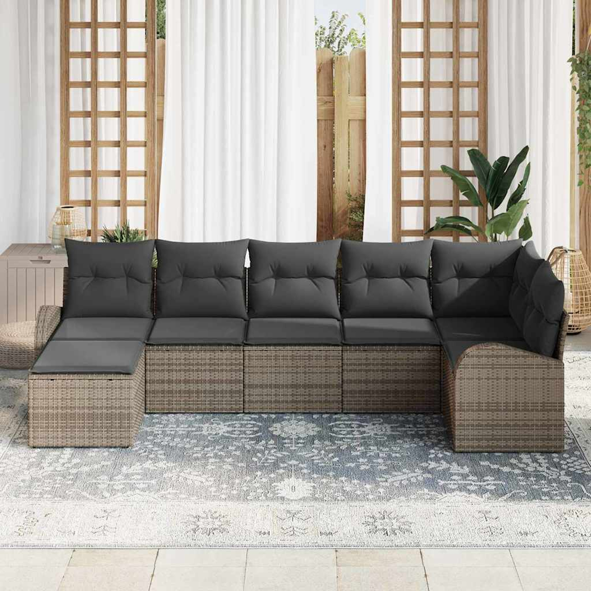 Ein graues Outdoor-Ecksofa mit Kissen steht auf einem gemusterten Teppich. Dahinter sind weiße Vorhänge und Holzgitter.