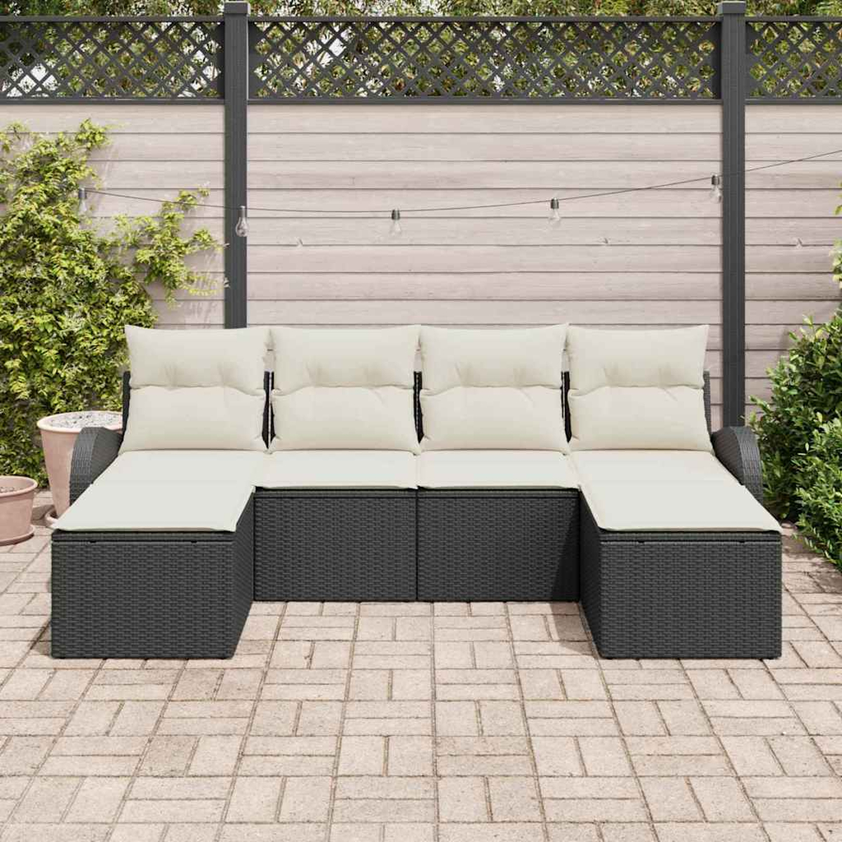 Ein dunkles Outdoor-Sofa aus Korbgeflecht mit cremefarbenen Kissen steht auf einer Backstein-Terrasse. Im Hintergrund befinden sich ein Holzzaun und Pflanzen.