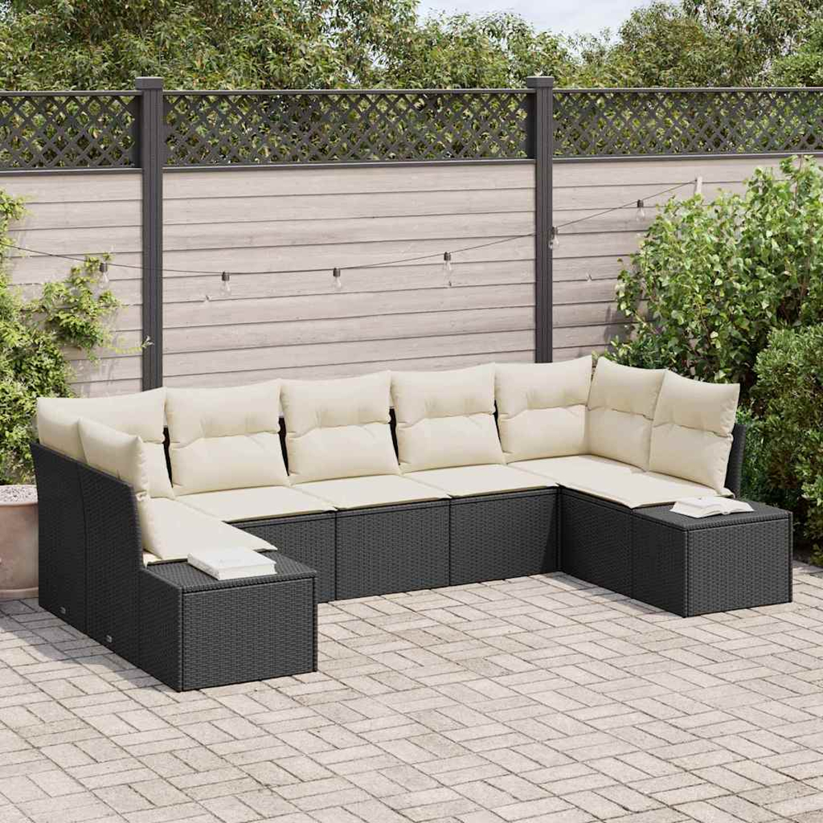 Ein schwarzes Rattan-Ecksofa mit cremefarbenen Kissen steht auf einer Ziegelterrasse. Ein Holzzaun und Grünzeug befinden sich im Hintergrund.