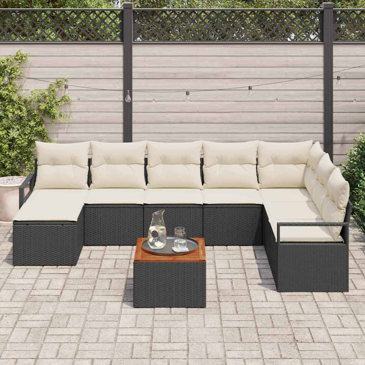 Outdoor-Ecksofa mit cremefarbenen Kissen und schwarzem Korbgeflecht. Ein Holztisch trägt eine Wasserkaraffe. Auf einer Terrasse mit Zaun.