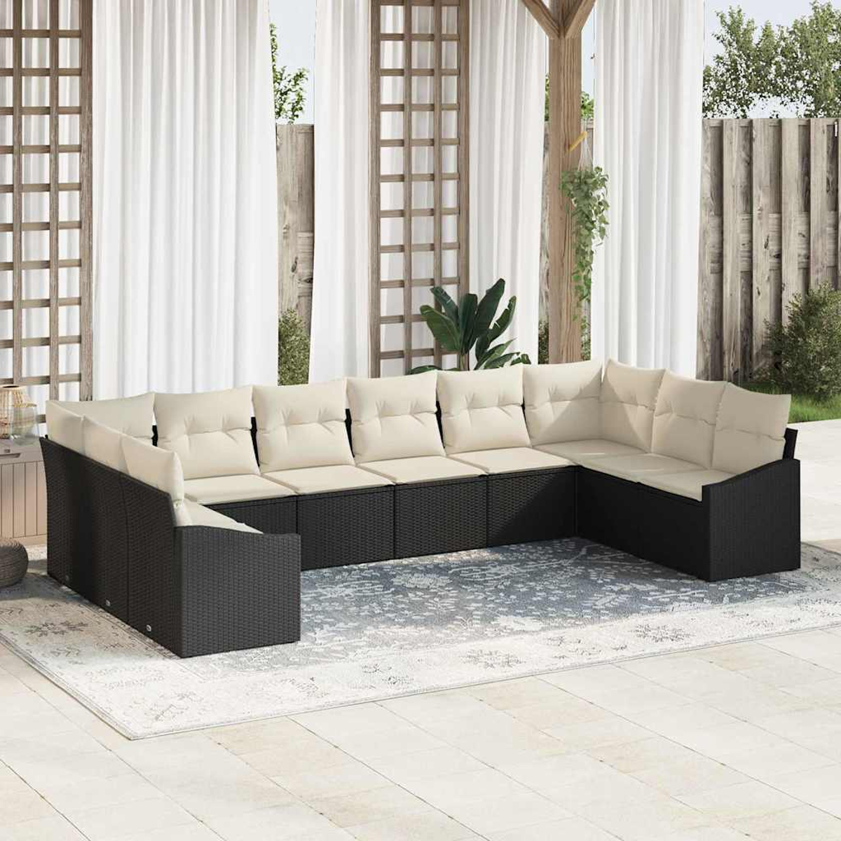 Ein schwarzes Korbsessel-Sofa mit cremefarbenen Kissen steht auf einem gemusterten Teppich. Weiße Vorhänge und Holzstruktur im Hintergrund.