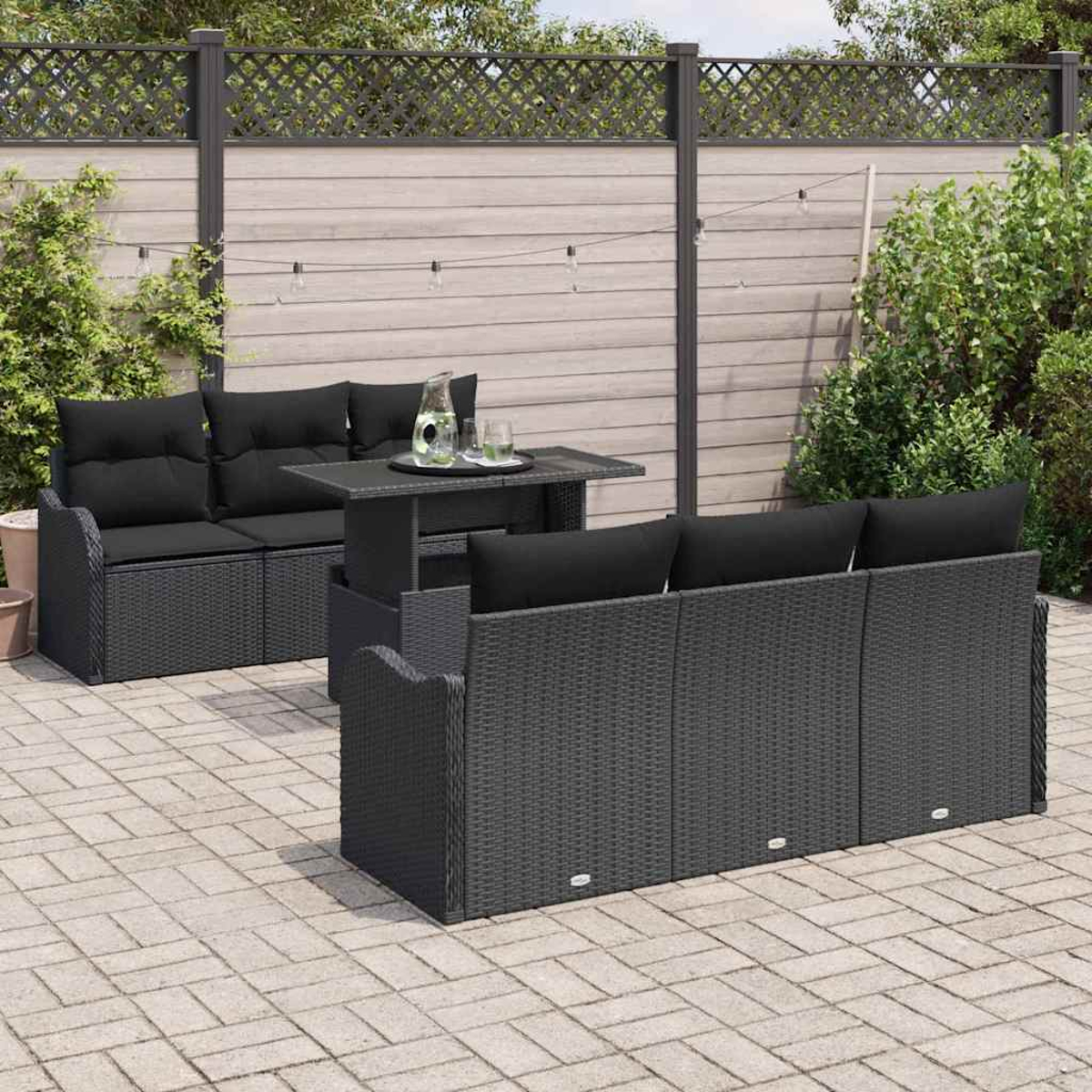 Terrassenset im Freien mit schwarzen Kissen und einem Tisch. Graue Korbmöbel auf einer gepflasterten Terrasse mit Holzzaun im Hintergrund.