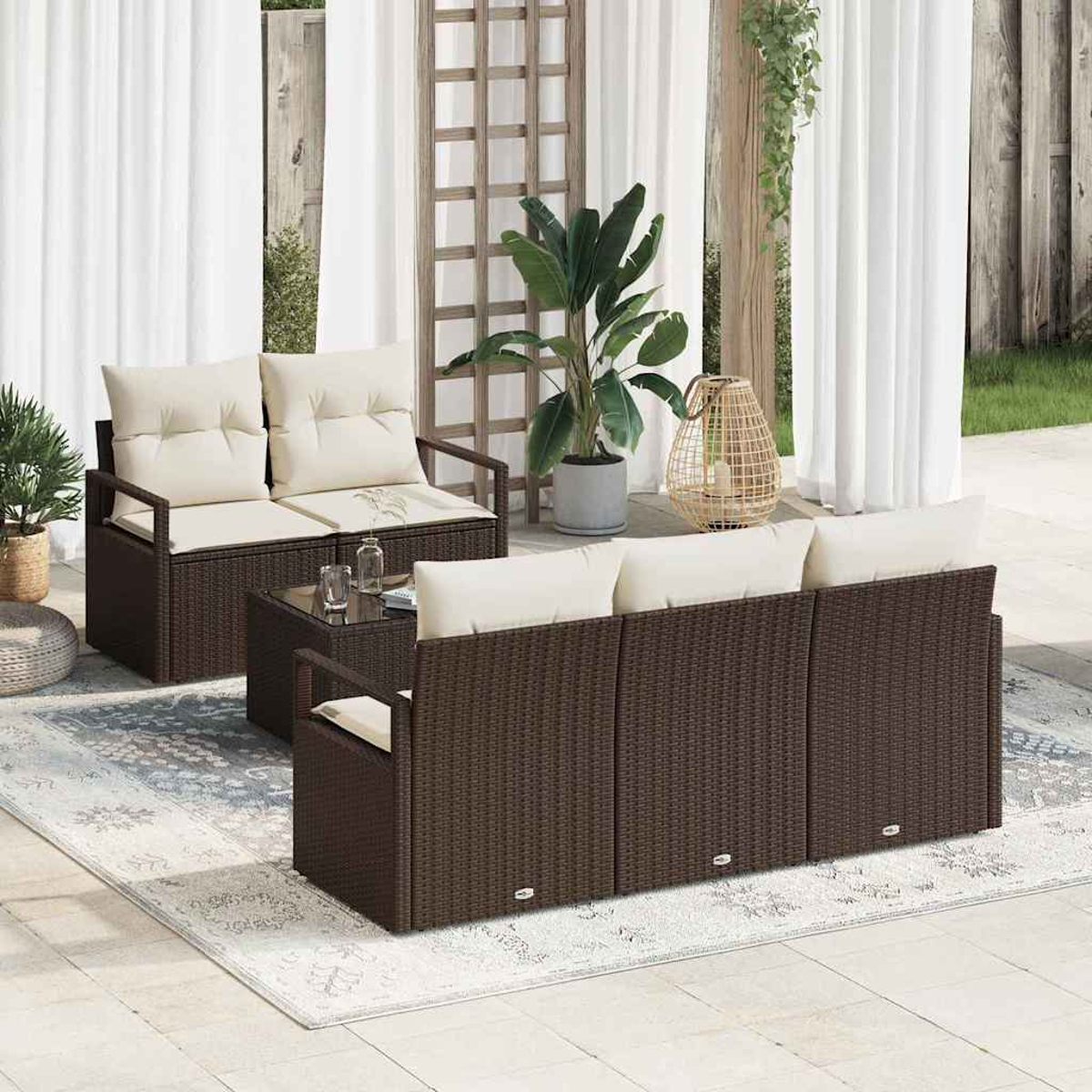 Gartenmöbel-Set mit braunen Korbmöbeln, cremefarbenen Kissen, Glastisch, Teppich. Hintergrund: Pflanzen, Vorhänge und Holzspaliere.