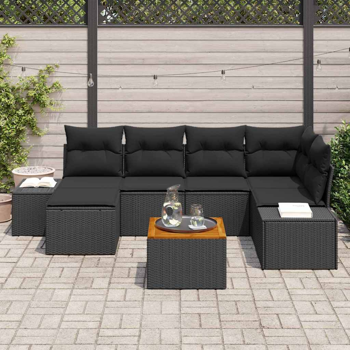 Gartencouch mit schwarzen Kissen, Holztisch mit Tablett und Backstein-Patio. Hintergrund: Holzzaun und Pflanzen.