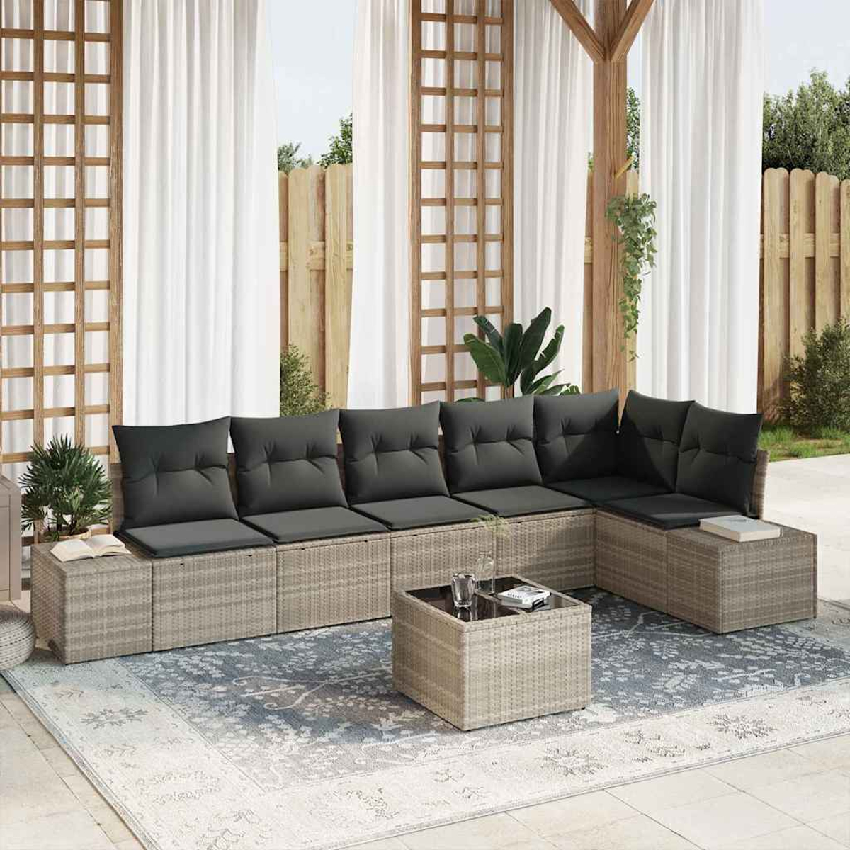 Außenbereich mit grauen Kissen, einem quadratischen Couchtisch und einem gemusterten Teppich. Weiße Vorhänge und Holzkonstruktionen im Hintergrund.