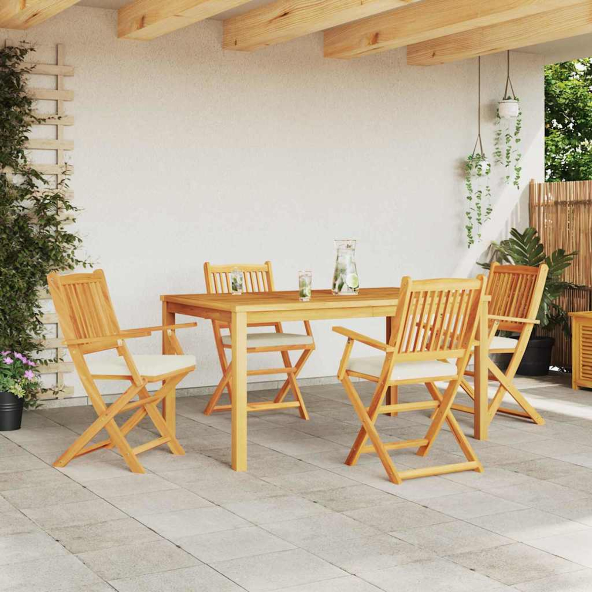 Eine Terrasse mit einem Holztisch und vier Stühlen. Auf dem Tisch stehen eine Wasserkaraffe und Gläser. Pflanzen und ein Spalier sind sichtbar.