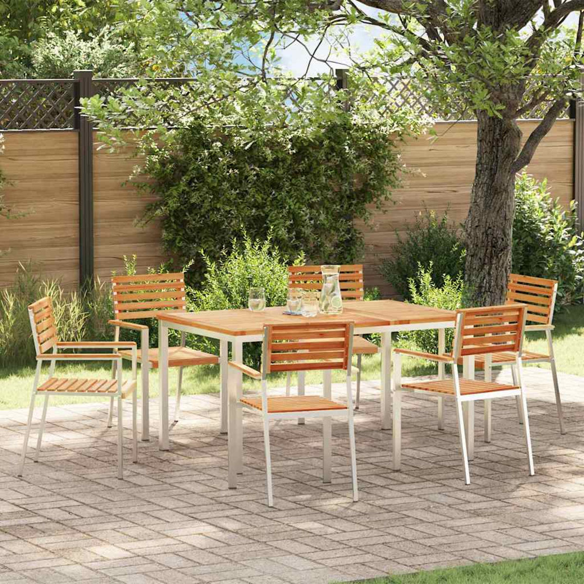 Juego de comedor al aire libre en un patio de ladrillos. Una mesa y sillas de madera están dispuestas. Follaje verde y una valla de madera al fondo.