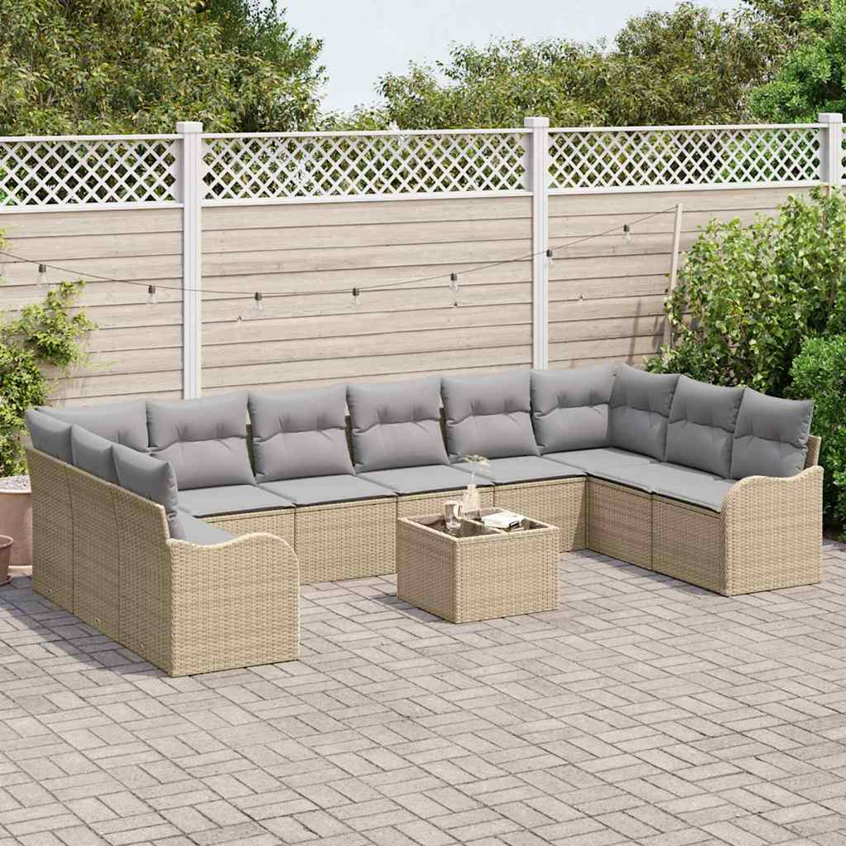 Ein beiges Korbsessel-Ecksofa mit grauen Kissen steht auf einer Terrasse. Ein passender quadratischer Tisch hat ein Glas. Dahinter ist ein weißer Zaun.