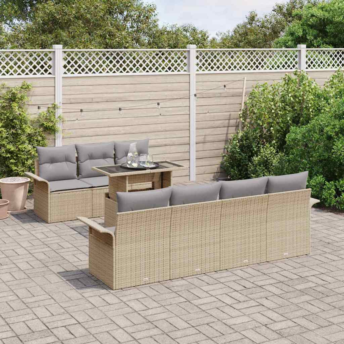 Set patio esterno con mobili in vimini beige e cuscini grigi, un tavolo con bevande e una recinzione bianca sullo sfondo.