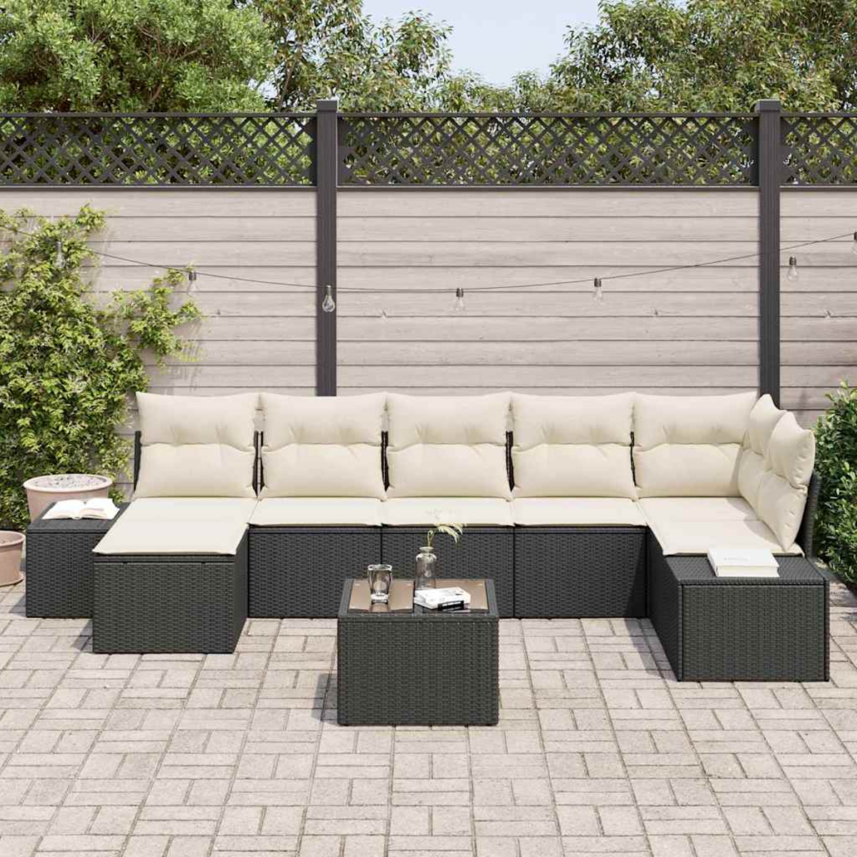 Outdoor-Ecksofa mit cremefarbenen Kissen, kleiner Tisch mit Glas und Büchern. Terrasse mit Ziegelpflaster, Zaun und Grünflächen im Hintergrund.