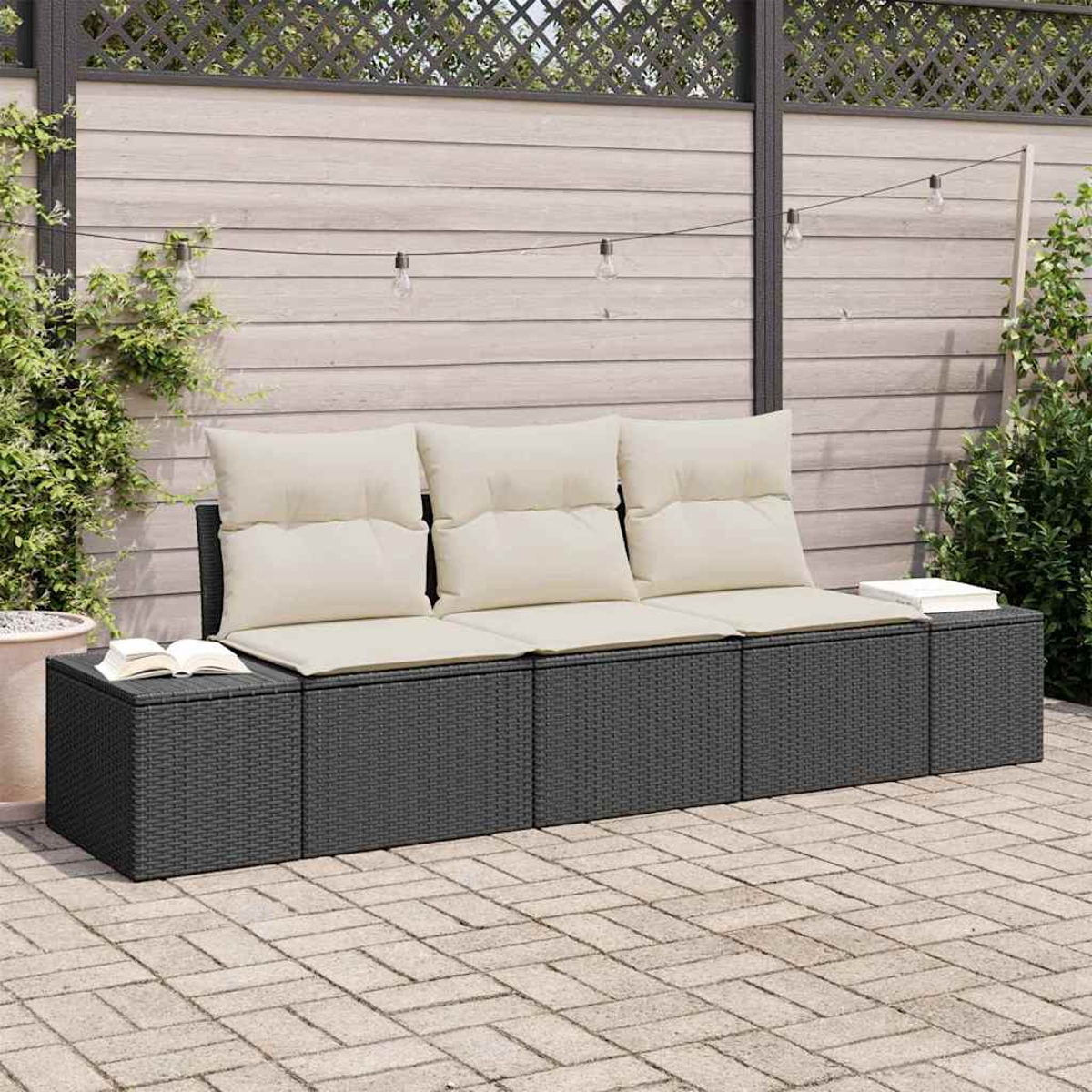 Outdoor-Sitzgelegenheit mit Kissen, Büchern und Lichterketten. Geflochtenes graues Sofa auf einer Steinterrasse, Holzzaun im Hintergrund.
