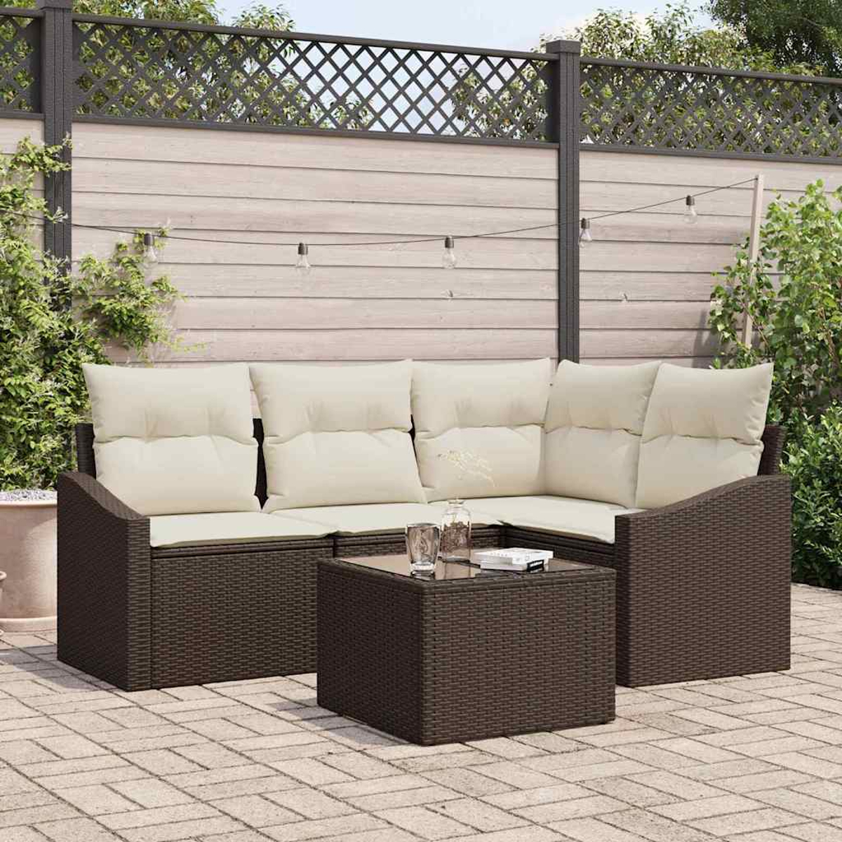 Garten-Set mit braunem Korbmöbel, beigen Kissen und kleinem Tisch. Hintergrund: Zaun, Pflanzen und Lichterkette.