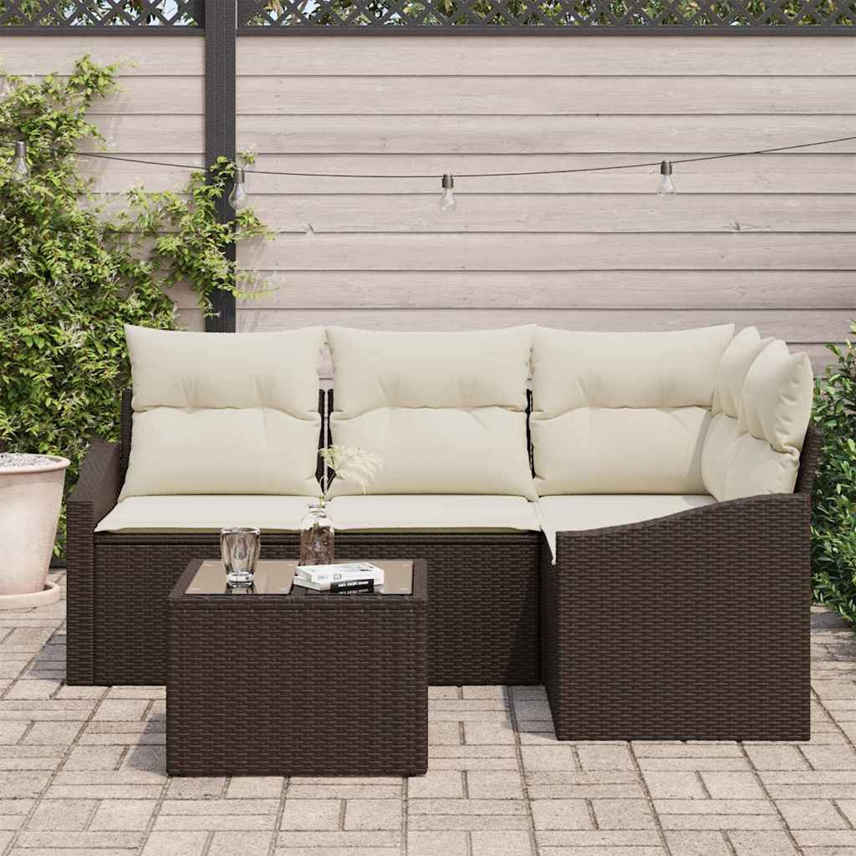 Braunes Outdoor-Ecksofa aus Korbgeflecht mit cremefarbenen Kissen und einem kleinen Tisch in einer Terrasse mit Holzzaun.