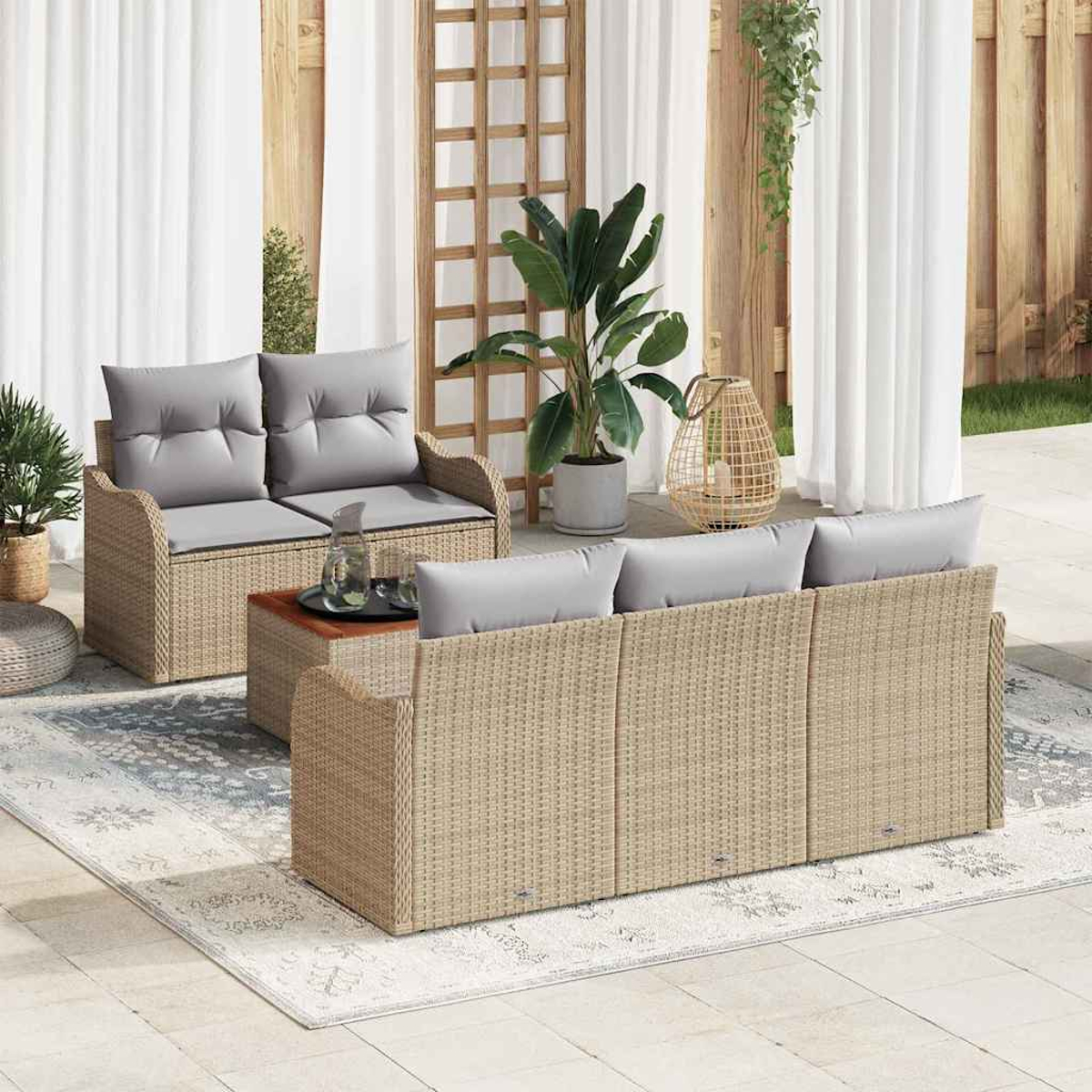 Juego de patio exterior con muebles tejidos beige, cojines grises, mesa de madera y alfombra estampada. Plantas y cortinas blancas al fondo.