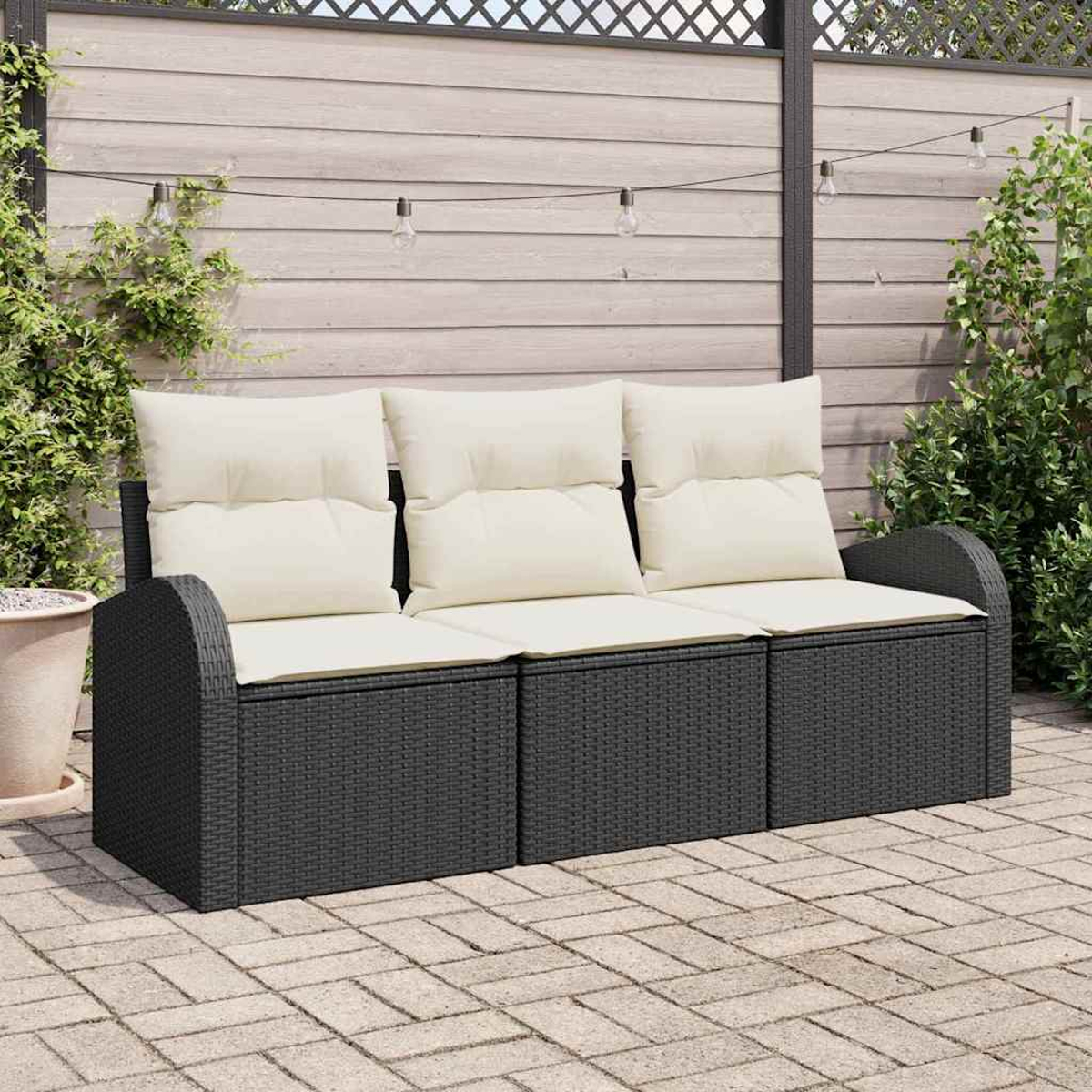 Ein schwarzes Outdoor-Sofa aus Korbgeflecht mit cremefarbenen Kissen steht auf einer Backstein-Terrasse. Im Hintergrund ein Holzzaun und Grün.