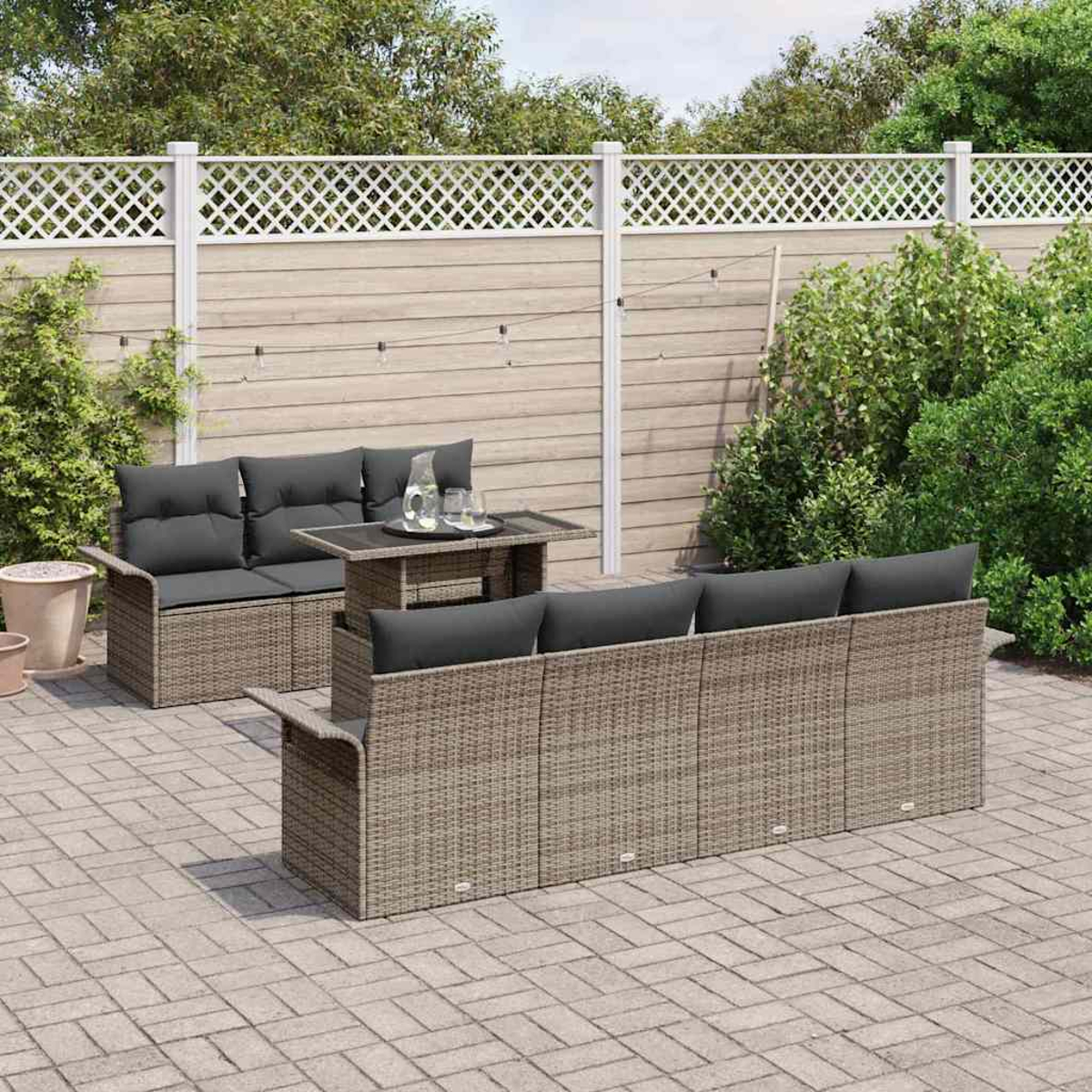 Set da patio all'aperto con mobili in vimini, cuscini grigi e tavolo con piano in vetro. Dietro, una recinzione con verde.