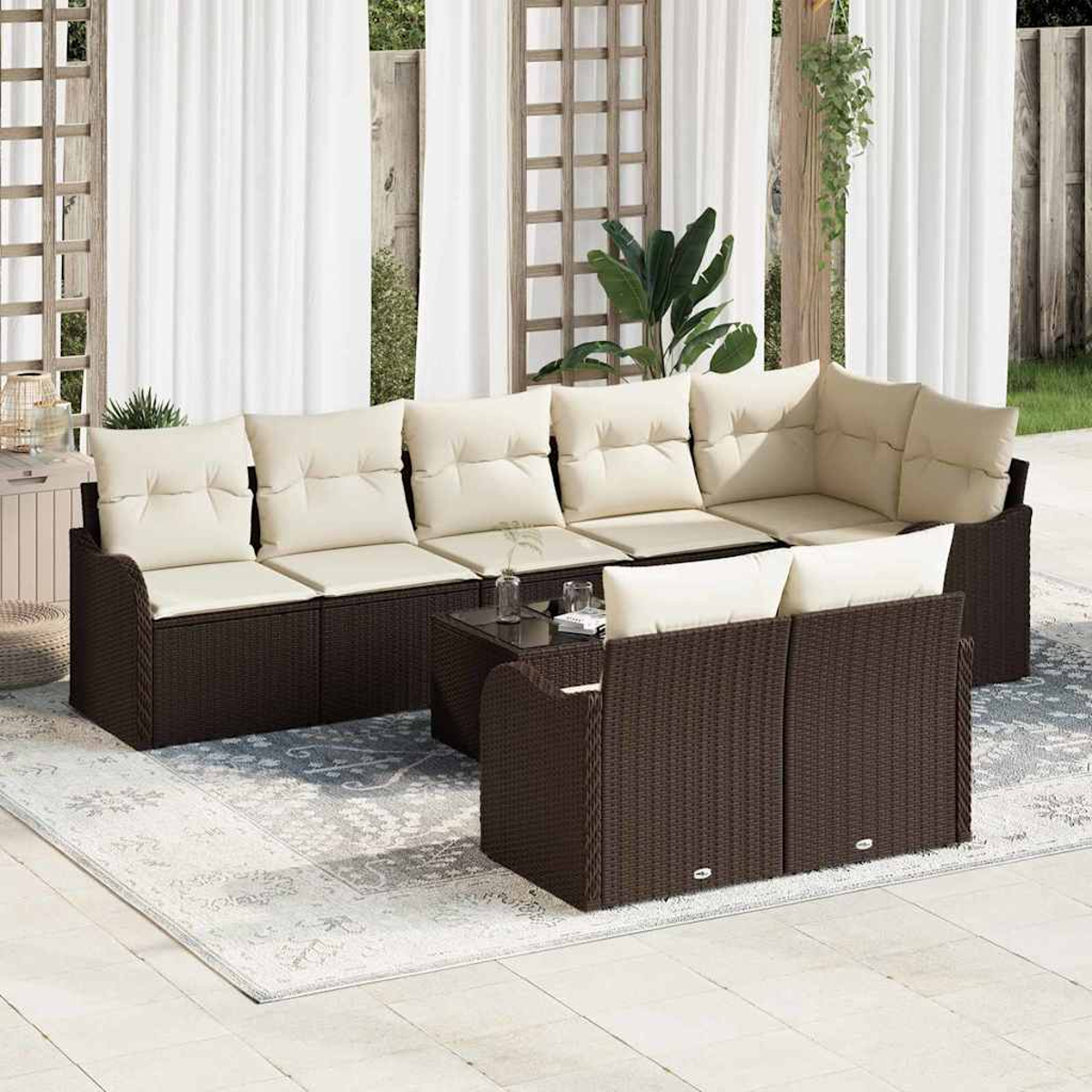 Sofá seccional de mimbre marrón para exteriores con cojines crema, mesa pequeña y alfombra en un patio con cortinas blancas.